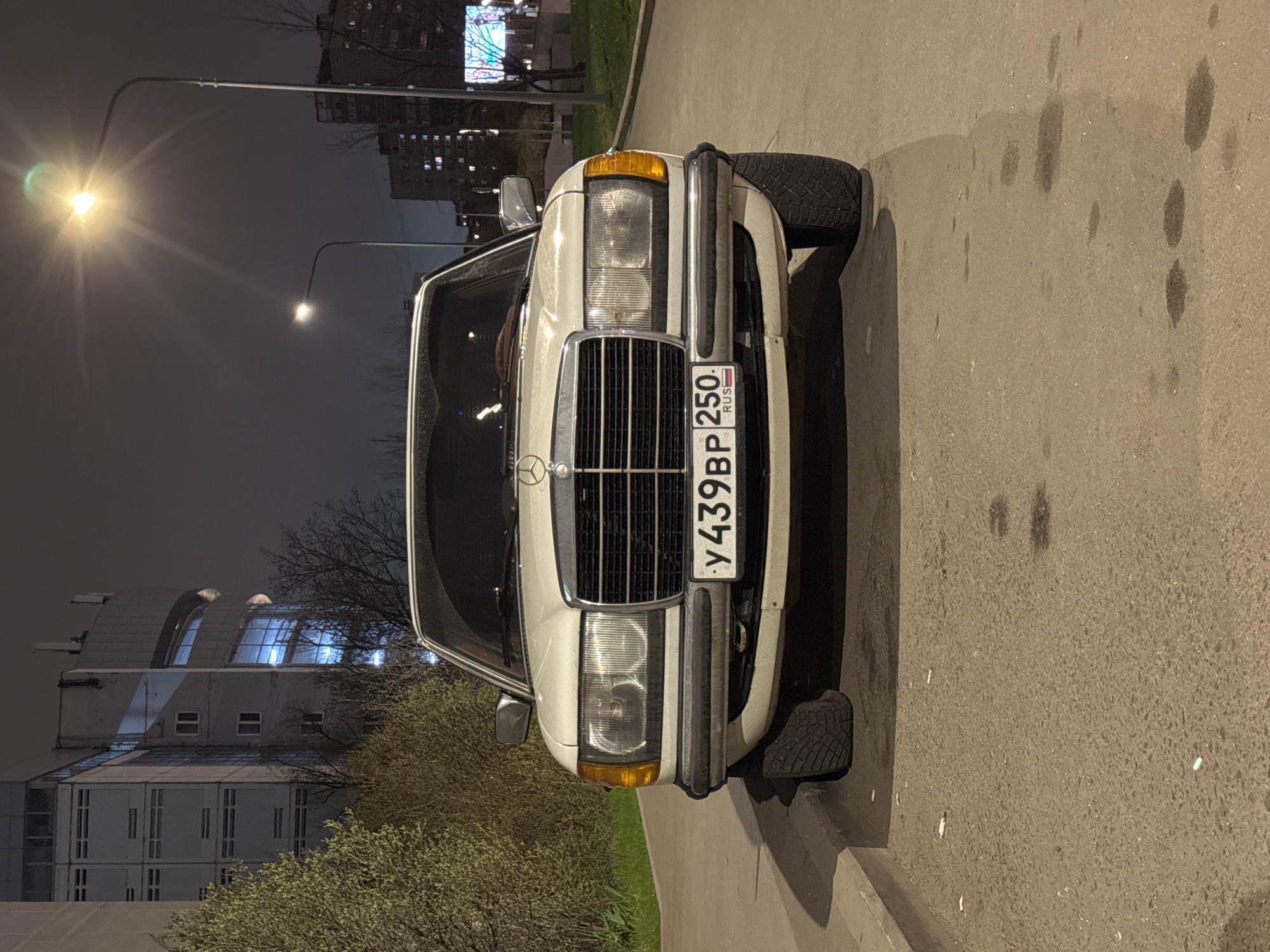 The image shows a white Mercedes-Benz car parked on a street in Moscow, Russia, at night. The car's license plate is visible, reading "У439ВР 250 RUS." A street lamp illuminates the scene. In the background are buildings, including one with distinctive architectural features, potentially a research or industrial facility. The surroundings are urban, with trees and pavement visible. There are no people in the immediate foreground. Oil or some other liquid stains are visible on the pavement.