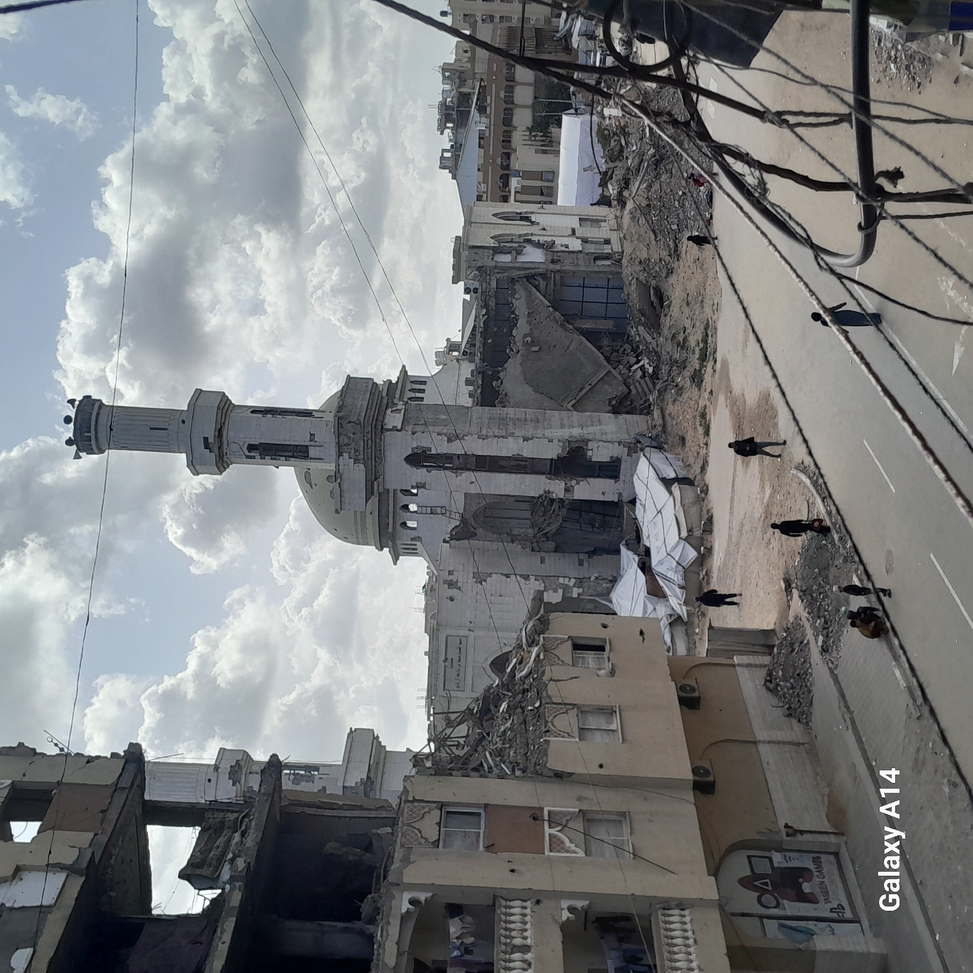 The image depicts a city street in Unknown, Unknown, marked by significant destruction. In the foreground, a multi-story building exhibits severe structural damage. A mosque, featuring a minaret and dome, stands partially destroyed in the middle ground. Debris is scattered along the street, where several individuals are walking. Utility wires are visible overhead, adding to the chaotic scene. The sky is filled with clouds.