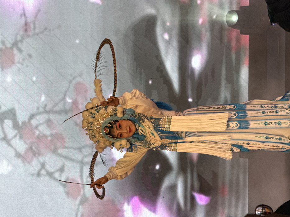 A single individual is depicted center-frame on a stage in Yuzhong District, China. The subject faces forward with a direct gaze, hands raised symmetrically above shoulder height, each grasping the base of a long, curved brown feather extending laterally from an ornate headdress. The headdress is composed of white and gold elements, faux pearls, and a prominent central red gemstone. Facial features are obscured by heavy theatrical makeup, including a white base, rouged cheeks, defined red lips, and exaggerated eye makeup. The costume is a floor-length white and blue garment, featuring intricate embroidery and multiple hanging white tassels with blue bead accents. The background is a large LED screen displaying a blurred image of pink blossoms against a gradient of light grey and purple hues. A stage light is visible in the lower right, and elements of an audience or stage equipment are partially visible in the lower left.