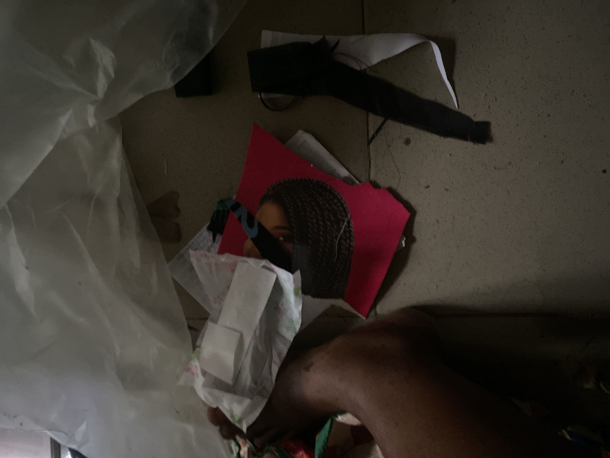 A person's left foot and leg are visible on the left side of the image, resting on a dirt or concrete floor. The person's skin tone is dark. Scattered around the foot are various items including what appears to be used feminine hygiene products, a torn piece of red paper with an image of a woman's braided hair, and a black ribbon tied with red thread. The scene is dimly lit, suggesting it may be indoors or at night. The overall impression is one of disarray and possible neglect. There is no specific information in the image to indicate the location in the city of Jauro Yinu, Nigeria, beyond the general appearance of the floor and the discarded items which are not culturally specific.