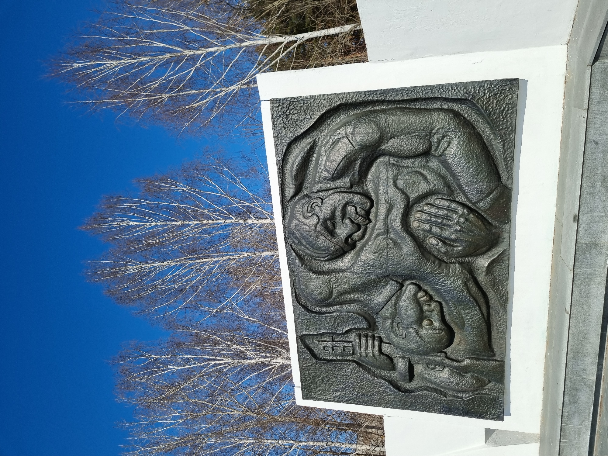 A dark, textured bas-relief sculpture, identified as the "Mother of a Soldier" monument, is centrally positioned on a white, angular wall in the Park of Victory, Chaikovsky, Russia. The sculpture depicts two stylized human figures: a larger, muscular figure with a pained expression embracing a smaller figure whose head rests against the larger one. The larger figure's left hand holds a stylized multi-story building, while its right hand supports the smaller figure. The white wall rests on dark grey stone steps or a plinth, partially visible at the bottom. Behind the monument, under a clear, deep blue sky, stands a dense line of tall, bare-branched deciduous trees, primarily birches with white bark. Sunlight illuminates the scene from the left.