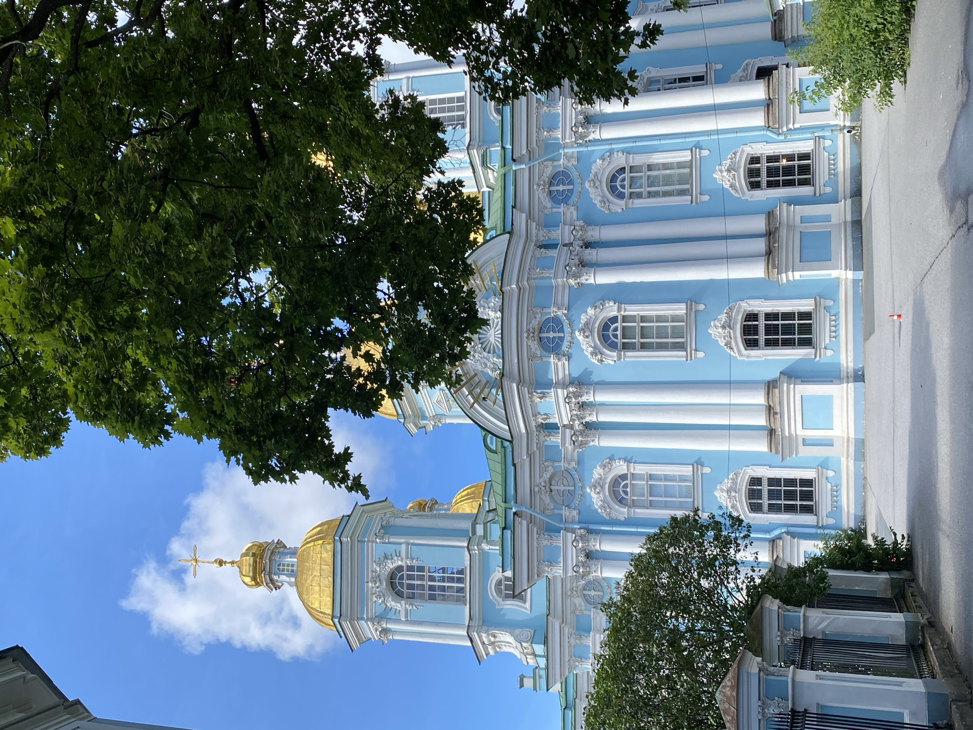 The image depicts the exterior of the Saint Nicholas Naval Cathedral in Saint Petersburg, Russia. The cathedral is a prominent blue building with white architectural details and ornate gold domes, one of which is visible at the top left, topped with a golden cross. The foreground shows a grey paved area and a black metal fence with blue and white decorative posts. Lush green trees with dense foliage partially obscure the view of the cathedral, framing the scene from the top right and left. The sky is blue with scattered white clouds. There are no people or visible actions in the scene.