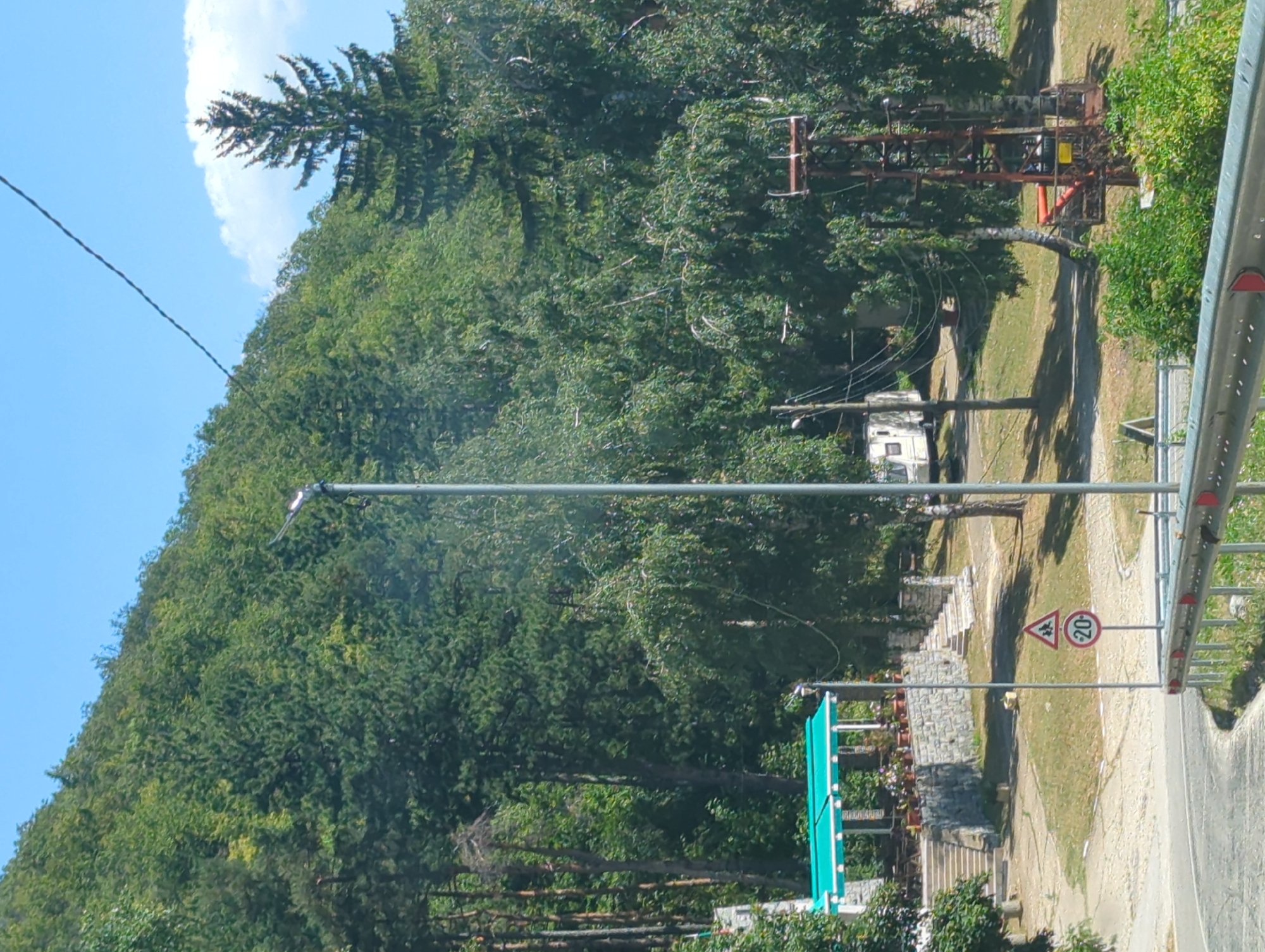 The image captures a scene in Vratsa, Bulgaria, on a clear, sunny day. A paved road curves through the foreground, bordered by a metal guardrail with red reflectors. Beyond the road, a grassy area is dotted with trees and a signpost displaying a children crossing symbol and a speed limit of 20. In the mid-ground, a stone building with a green awning and potted plants is visible, accessible by stone stairs. Further back, a white camper van is parked near a utility pole. To the right, a rusty, industrial-looking structure stands amidst the trees. The background is dominated by a densely wooded hillside, with a prominent tall pine tree in the upper right corner. The sky is a clear blue with a few scattered white clouds.
