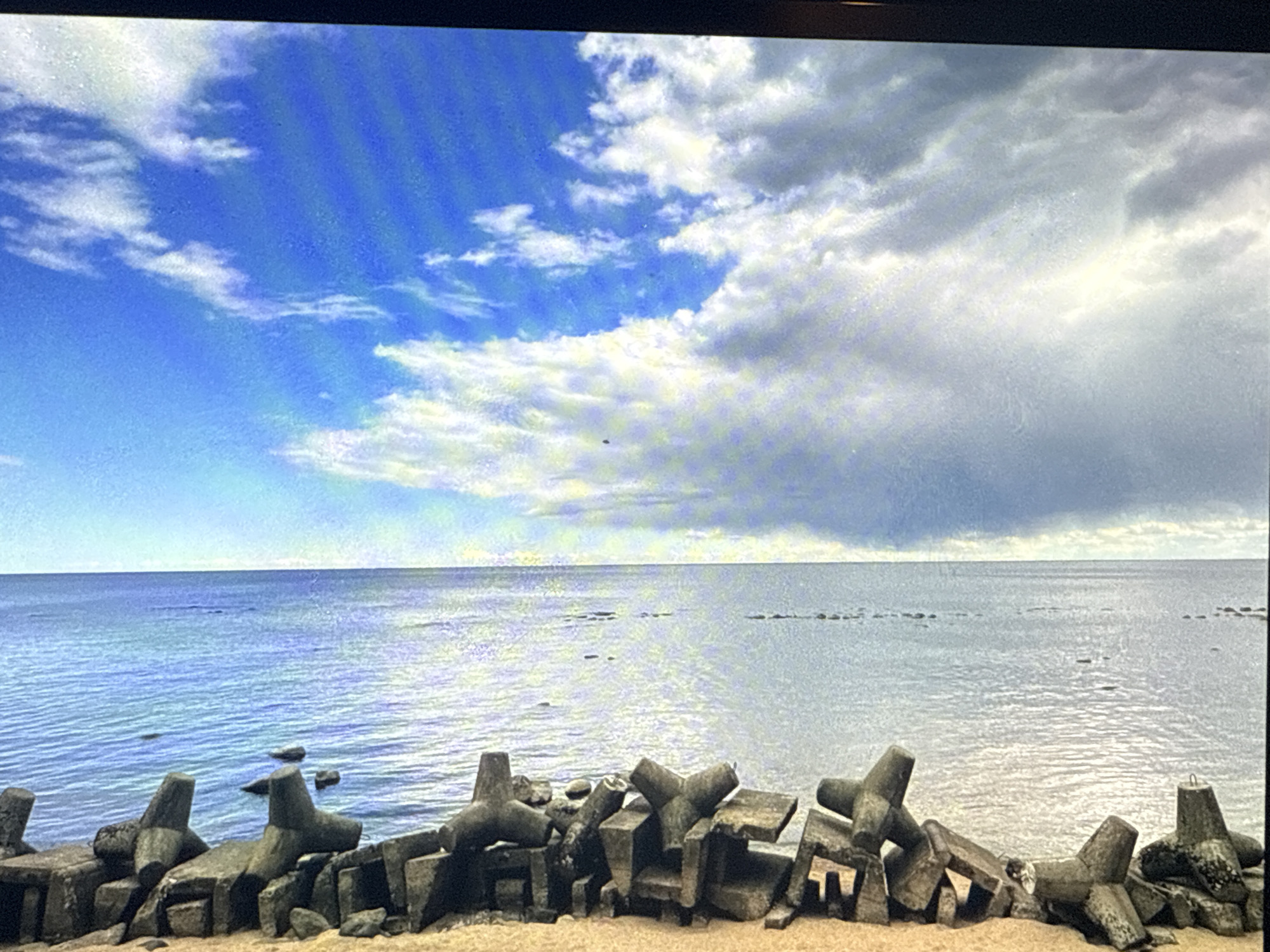 The image depicts a seascape. In the foreground, there is a row of concrete tetrapods on a sandy surface. Beyond that is the ocean, with water stretching towards the horizon. The sky is partly cloudy, with blue visible between the white clouds. The light reflects on the surface of the ocean.
There is no indication of the location being Moscow, Russia in the image.