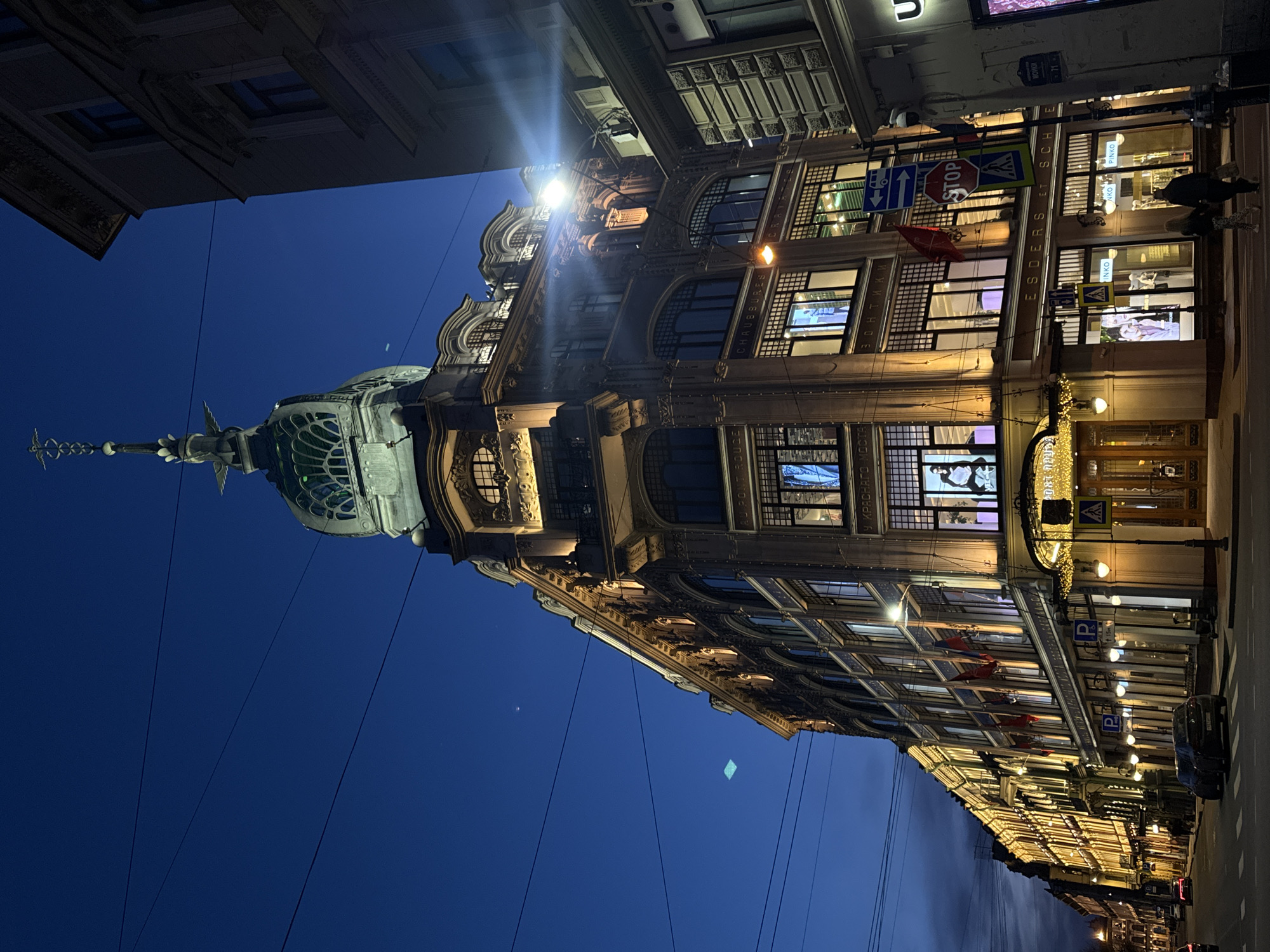 The image captures a nighttime scene in Saint Petersburg, Russia, featuring the iconic Au Pont Rouge department store. The architectural structure of the building is emphasized by the illumination, highlighting its ornate facade, dome, and spire. The store fronts are lit, displaying merchandise and mannequins in the windows. The street scene includes traffic signs, street lights, and parked vehicles, with a couple of pedestrians walking along the sidewalk. The sky is a deep blue, transitioning to a darker shade near the horizon. The composition includes parts of adjacent buildings, indicating an urban setting.