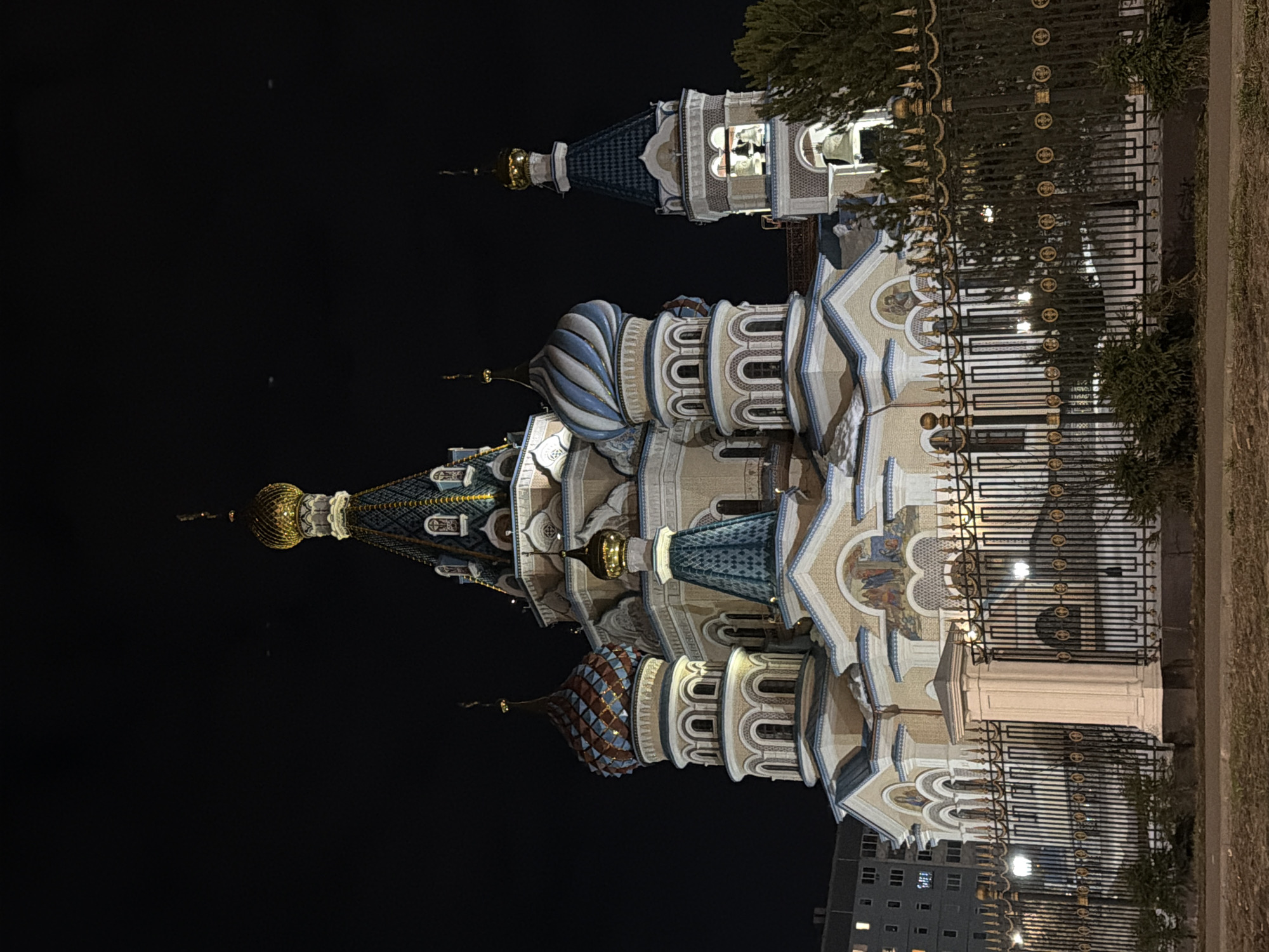 The image captures the architectural marvel of St. Michael's Cathedral in Izhevsk, Russia, illuminated against the night sky. The cathedral's intricate details are prominent: its multi-tiered structure featuring numerous domes topped with crosses, varying in color and design from golden to blue-and-white striped.
The building's façade is ornate, showcasing arched windows, mosaic decorations, and intricate stonework. The cathedral is partially obscured by a decorative wrought-iron fence and a small amount of greenery, suggesting a boundary between the building and public space. In the background, a few residential buildings and some additional greenery can be seen to the right and bottom of the image. The sky is dark, providing a stark contrast that emphasizes the illuminated architecture.
