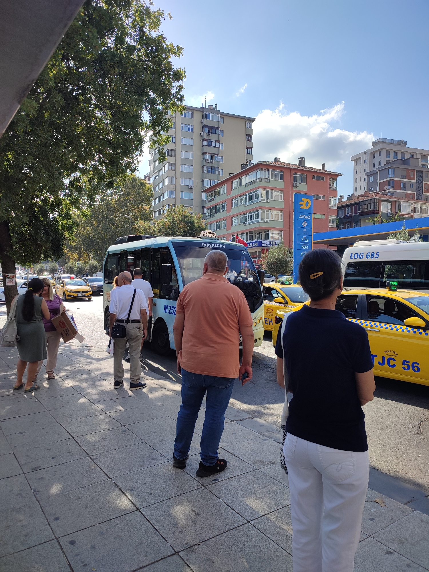 In Kadıköy, Turkey, a group of people are standing on a sidewalk next to a street. The scene is illuminated by sunlight, casting shadows on the gray pavement. A small blue bus with "M252" visible on its side is parked at the curb, and several individuals are either boarding or waiting to board. Behind the bus, yellow taxis are present, one of which has "TJC 56" on its rear. In the background, multi-story buildings line the street, including one with a prominent "AYGAZ" sign. Trees with green foliage are also visible, and the sky is partly cloudy. The people on the sidewalk are casually dressed, with some holding shopping bags. The overall impression is of a typical urban scene with public transportation and daily life unfolding.