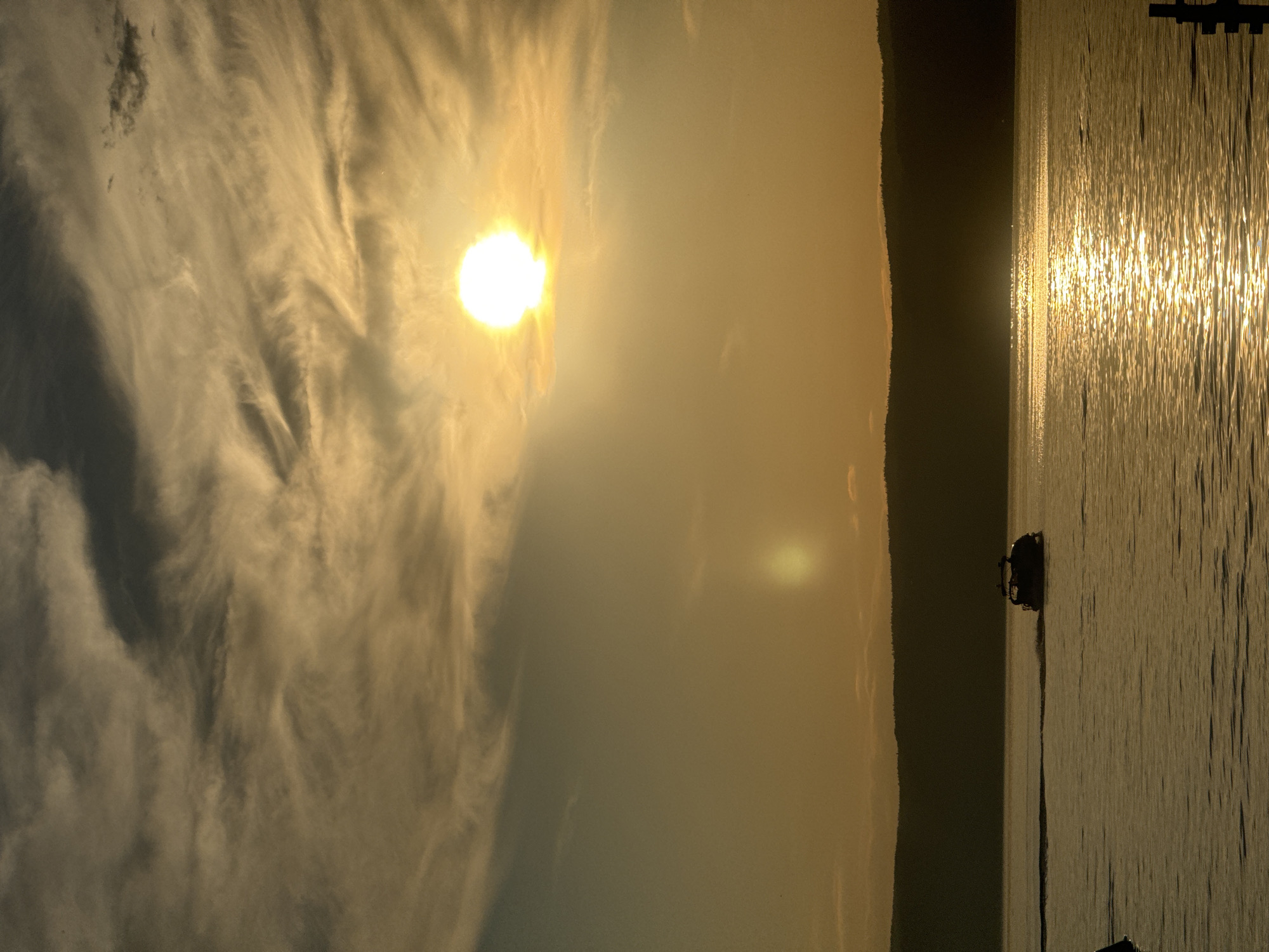 This is a vertical, eye-level, medium shot of a sunset over Lake Baikal, taken from the shore of Listvyanka, Russia. The sun, a bright white orb, is positioned in the upper right quadrant of the frame, partially obscured by wispy, grey and golden clouds that stretch across the sky. The water is calm and reflects the golden hues of the sunset, with shimmering streaks of light dancing on its surface. In the middle ground, a dark, silhouetted motorboat with an open cabin is moving from right to left, leaving a gentle wake behind it. The distant shoreline is a dark, undulating silhouette of trees and hills, also in shadow. In the foreground, on the left edge, a portion of a dark, blurred object, possibly a structure or another boat, is visible. On the right edge, a dark, vertical post, part of a pier or marker, juts out of the water. The overall mood is serene and tranquil.