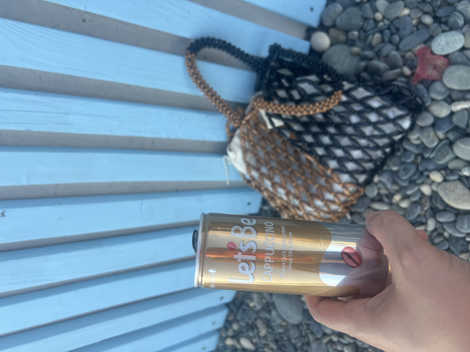 A person's hand holds a gold-colored aluminum can of "Let's Be CAPPUCCINO" with a silver band at the bottom. The can has the brand name in white and pink, a coffee bean graphic, and text that reads "Always Let's be together! Since 1991, Korea." Behind the can, a woven bag with black and tan handles and a black and tan mesh body is visible, partially filled with a white object. The background consists of a light blue wooden fence composed of vertical slats and a pebbled ground. The scene appears to be outdoors, possibly at a beach or waterfront, consistent with a location in Sokhumi, Abkhazia.