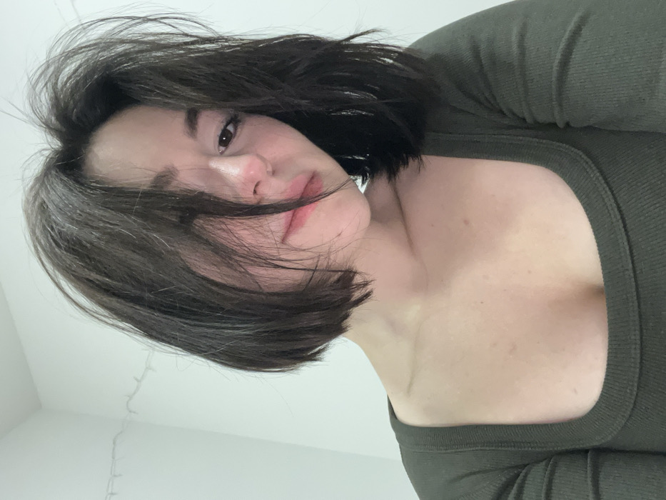 A young woman with dark, shoulder-length hair and fair skin looks downward at the camera. Her hair is somewhat tousled, with strands falling across her face. She has a neutral expression. The upper part of her torso is visible, clad in a form-fitting, long-sleeved top with a square neckline in an olive green color. The background is a plain white wall. The image is taken from a slightly angled, downward perspective.
