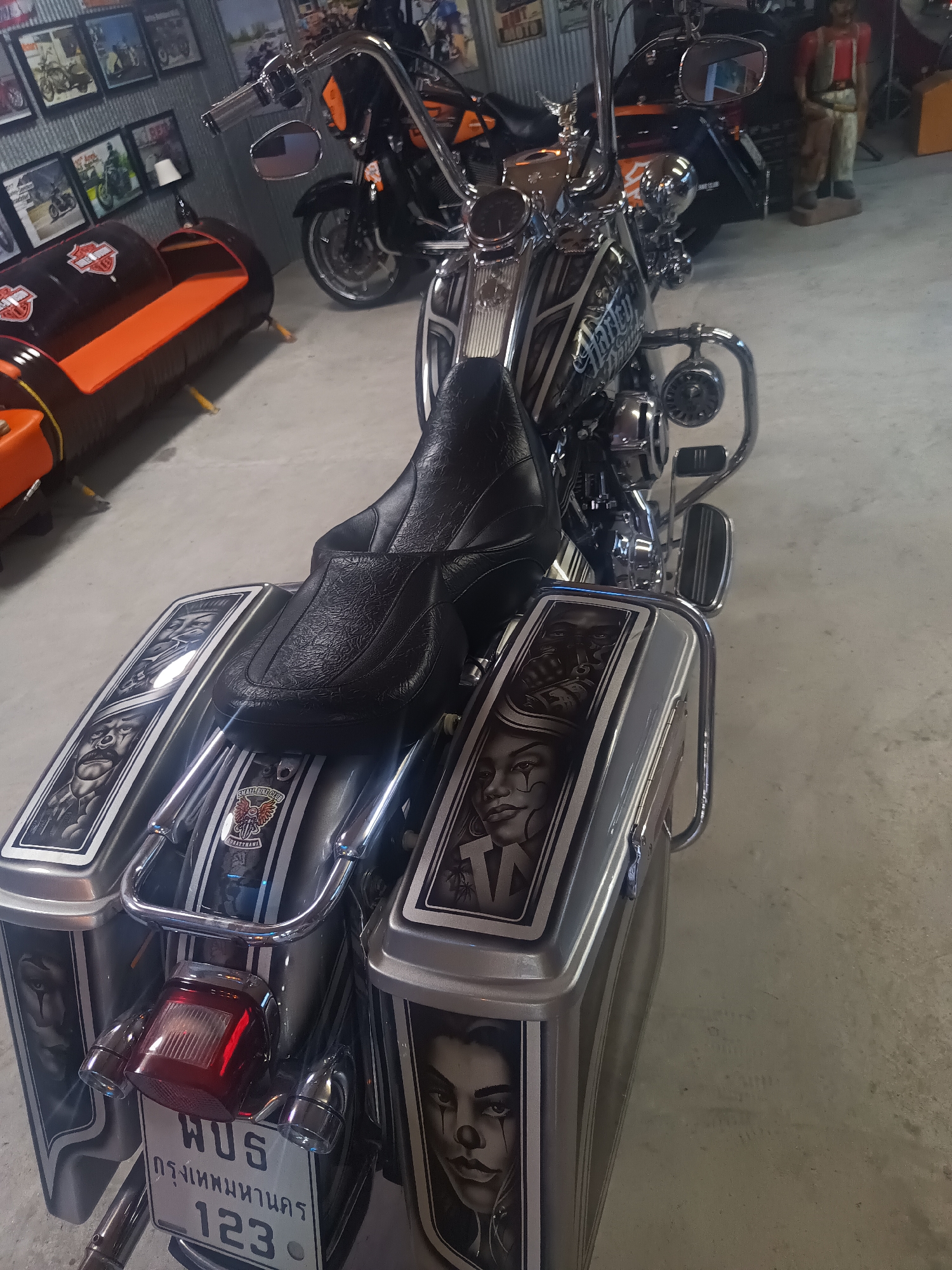 The image shows a Harley-Davidson motorcycle with custom artwork on its saddlebags. The artwork appears to be detailed, grayscale portraits of clowns. The motorcycle's license plate is partially visible and shows Thai script and numbers. The license plate includes the words "กรุงเทพมหานคร", which translates to Bangkok Metropolitan, and the number 123. The motorcycle is indoors, possibly in a showroom or garage. In the background, there is another motorcycle, a couch made of barrels, and wall decor featuring motorcycle-related images. The specific location is Surat Thani City Municipality, Thailand.