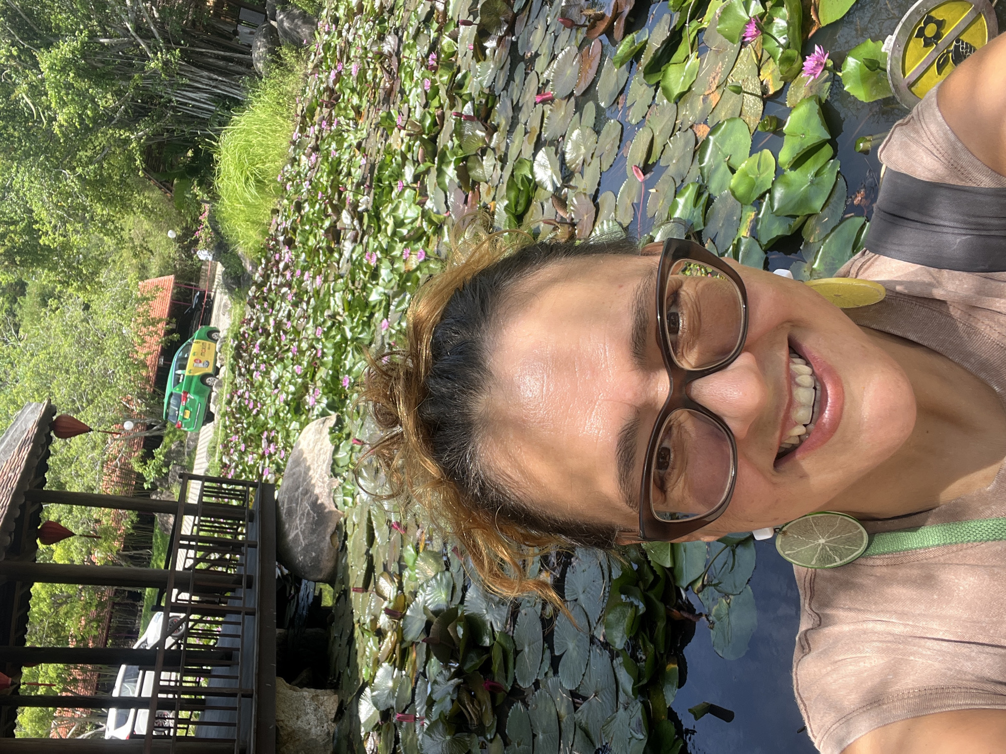 In Tay Nha Trang Ward, Vietnam, a woman with curly brown hair, wearing glasses and a light brown shirt, smiles at the camera in a selfie. She is adorned with lime slice earrings. Behind her, a pond is covered with numerous green lily pads, some with small pink lotus flowers. In the background, a wooden pavilion with red lanterns stands near a road where a green and yellow taxi is parked. Lush green trees and foliage surround the scene.