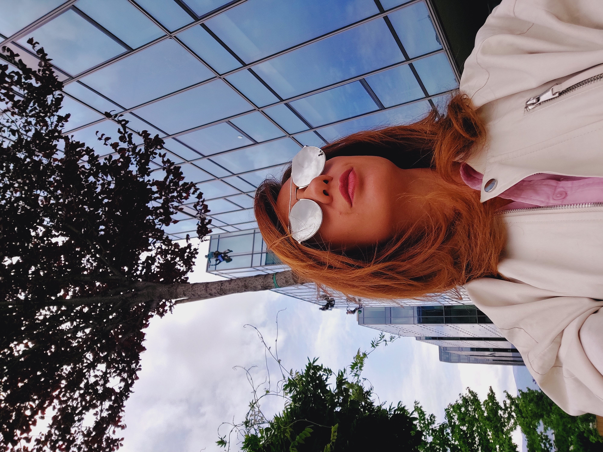 A woman with auburn hair and silver sunglasses is looking up at a modern glass-fronted building in Sofia, Bulgaria. She is wearing a light-colored jacket over a pink shirt. Several window cleaners in safety harnesses are visible on the side of the building, descending in separate groups. A tree with dark foliage is in the foreground to the left of the building. The sky is overcast.
