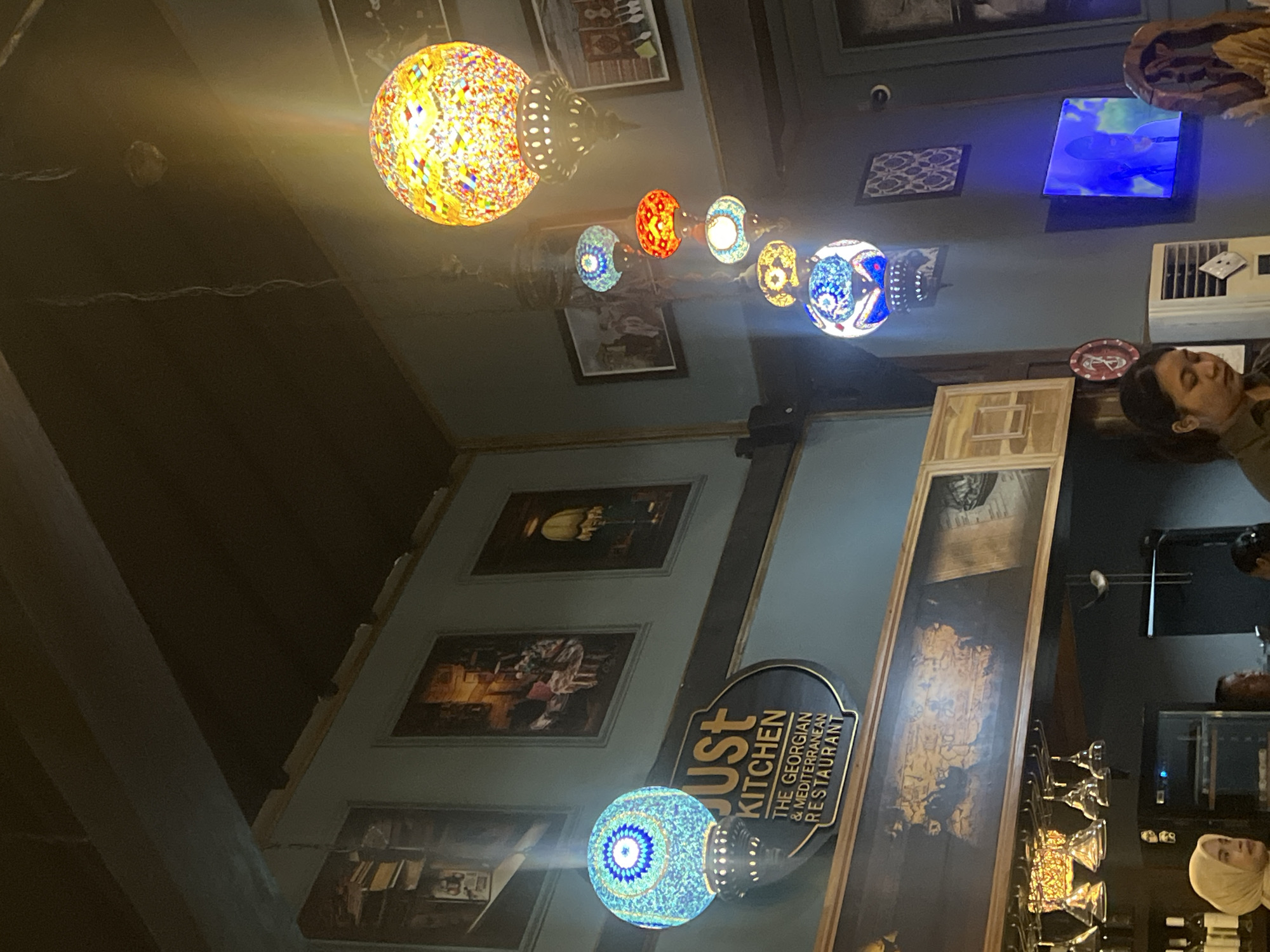 The image shows the interior of Just Kitchen, a Georgian and Mediterranean restaurant. A series of decorative lamps hang from the ceiling, featuring intricate glasswork. Paintings are mounted on the walls. A customer is present, visible in the background near an air conditioning unit. Wine glasses are partially visible. The location is North Kuta, Indonesia.