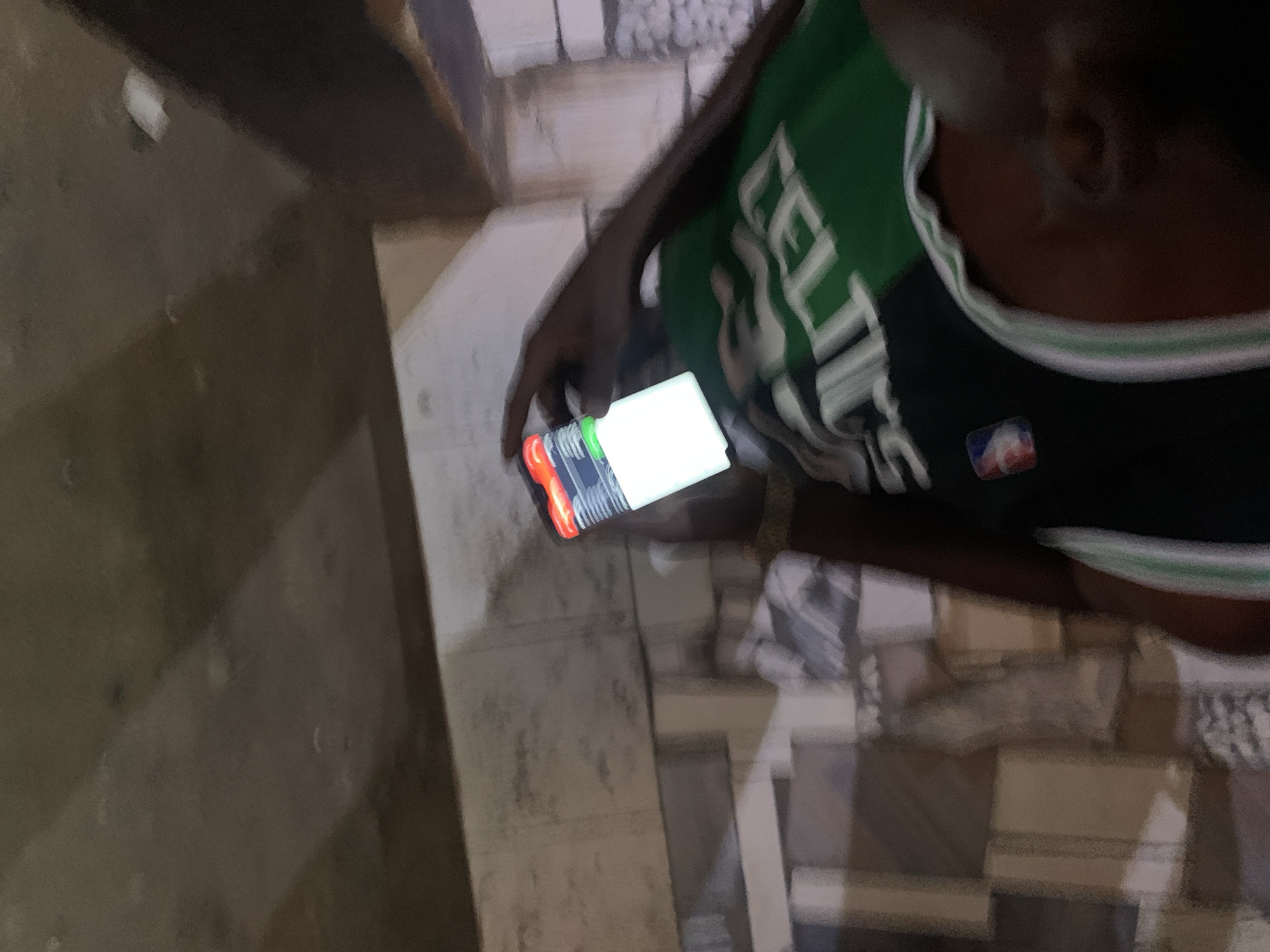 A person wearing a Boston Celtics jersey is holding a brightly lit mobile phone. The location appears to be in an urban area with brick or tiled flooring. The environment is dimly lit, suggesting it is indoors or it is night. The person is looking at the phone. The city of New Stadium, Nigeria is not definitively recognizable from the provided visual information.