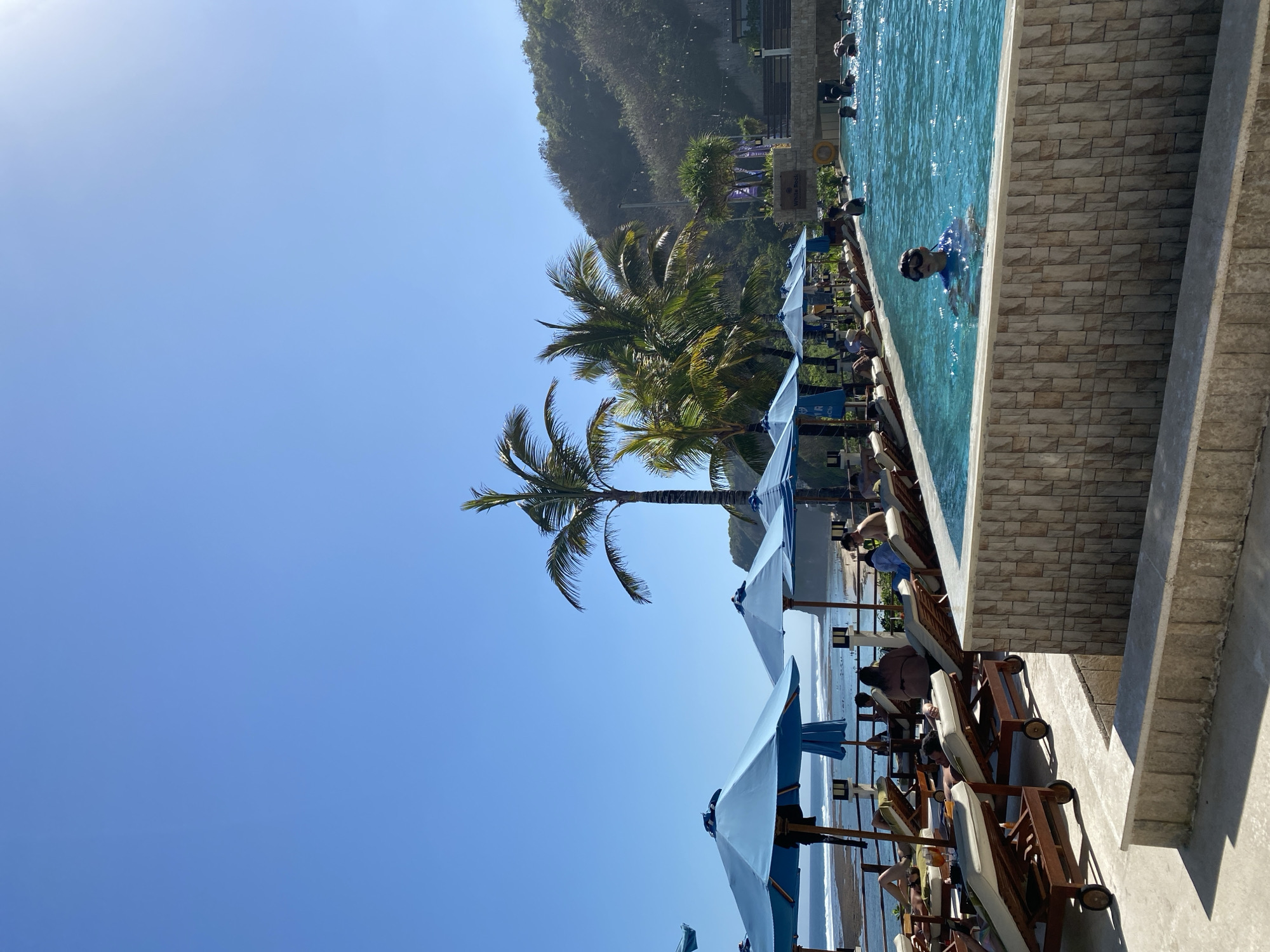 An exterior shot captures a resort pool area on a sunny day in Kuta Selatan, Indonesia. A large, bright blue swimming pool dominates the foreground, with a young boy wearing goggles and a blue shirt visible in the water. The pool is bordered by a textured stone wall and a concrete ledge. Beyond the pool, several lounge chairs with people relaxing are arranged under blue umbrellas. A wooden walkway runs along the edge of the pool, leading to the beach where waves are breaking. Palm trees and lush green hills rise in the background, indicating a tropical setting. The sky is a clear, vibrant blue, with a hint of the sun's glare on the right side of the image. The overall scene depicts a leisurely vacation atmosphere at a beachfront resort.