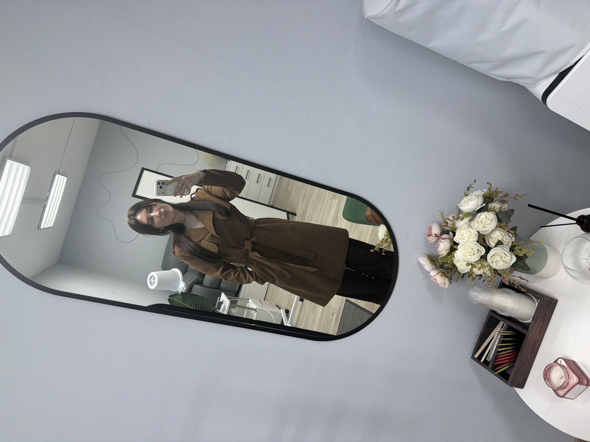 A person is taking a photo in a mirror in a room. The room contains florescent lighting, a framed picture, seating, a blower, wood-look flooring, and a light-colored wall. Next to the mirror is a table with a flower arrangement, glasses, and a wooden box. The box holds drink stirrers, and cups. A candle sits on the edge of the table. The location of the image is Perm, Russia.