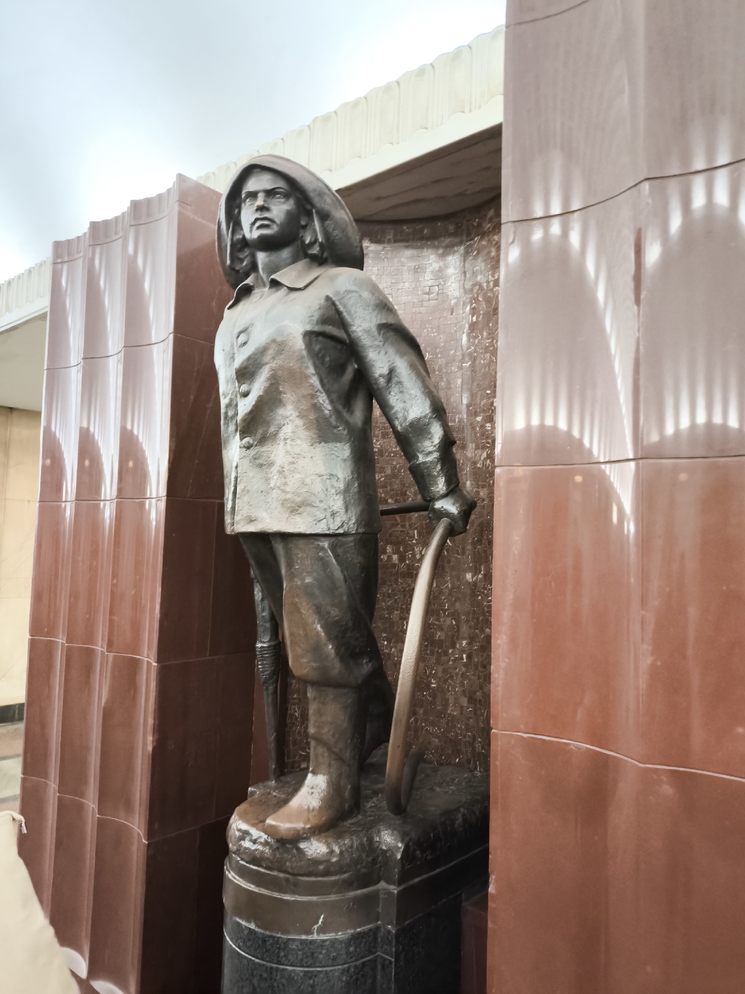 The image shows a bronze statue of a person in rain gear, standing on a tiered base made of black stone. The statue is situated in an alcove with walls made of alternating vertical panels of red and brown polished stone. The person is wearing a buttoned jacket and a hood. They are holding a long-handled tool in their right hand. The statue appears to be an outdoor monument located in a city. The location is Unknown, Unknown.