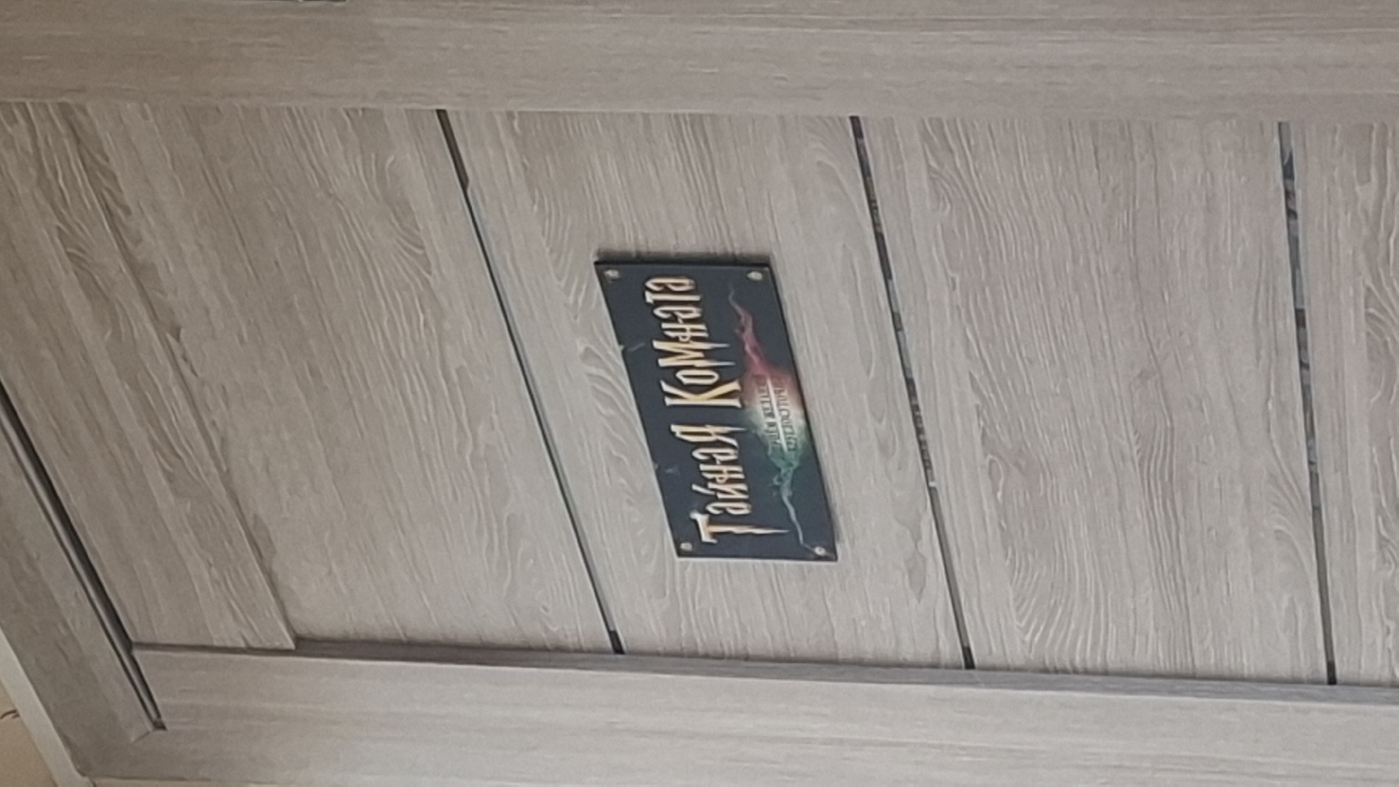 The image shows a close-up of a light-colored wooden door with a dark rectangular sign attached to it. The sign has the words "Тайная Комната" (Secret Room) written in gold, stylized lettering. Below this, there is smaller text that is difficult to read but appears to be an address or description. The door itself has horizontal paneling with a visible wood grain pattern. The lighting suggests an indoor environment. There are no people or animals visible in the image. The location in Shymkent, Kazakhstan cannot be determined from the image itself.
