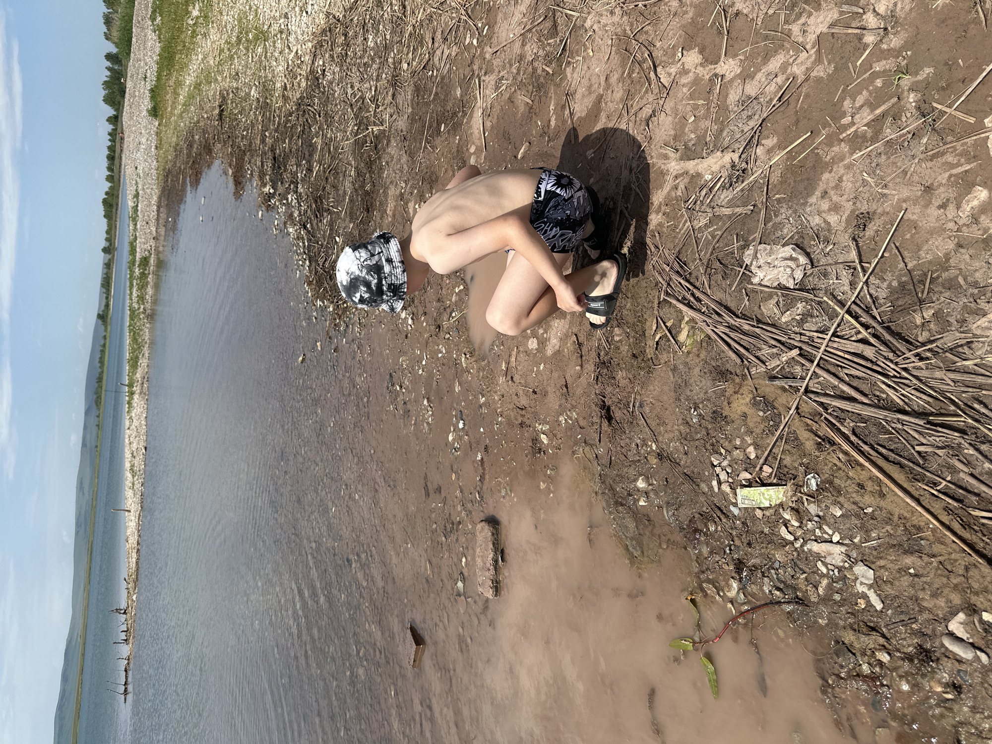 In Novonikolaevka, Russia, a young boy wearing a black and white patterned bucket hat and black and white patterned swim trunks crouches by the murky water's edge. He is wearing black sandals. The boy is facing away from the camera, looking at the water where two rectangular objects, possibly bricks, are partially submerged. The ground is muddy and littered with small stones and dry reeds. The background shows a wide expanse of water under a partly cloudy blue sky, with hills and sparse trees in the distance.