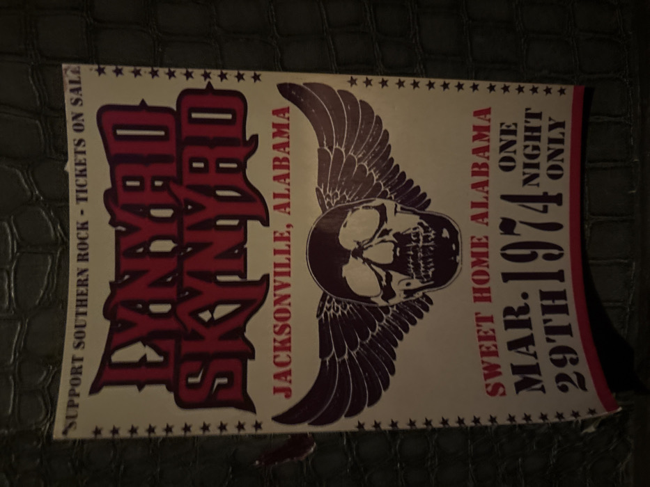 A vintage-style Lynyrd Skynyrd concert poster is depicted, observed in Mexico City, Mexico. The rectangular poster is predominantly off-white with red and maroon typography and graphics, presented against a dark, textured background resembling reptile skin. A border of small red stars frames the poster's perimeter. The top section of the poster displays the text "SUPPORT SOUTHERN ROCK - TICKETS ON SALE." Below this, "LYNYRD SKYNYRD" is prominently featured in large, stylized red block letters with a darker outline. Directly beneath the band name, "JACKSONVILLE, ALABAMA" is printed in red. The central graphic consists of a maroon stylized human skull with outstretched feathered wings. Below the graphic, the text "SWEET HOME ALABAMA" is present in red, followed by "MAR. 29TH 1974" in large, stencil-like dark red font. To the right of the date, "ONE NIGHT ONLY" is vertically aligned in smaller red text. The poster exhibits minor surface imperfections, including subtle creasing and a slight curl along its bottom edge. No individuals or specific actions are visible within the frame.
