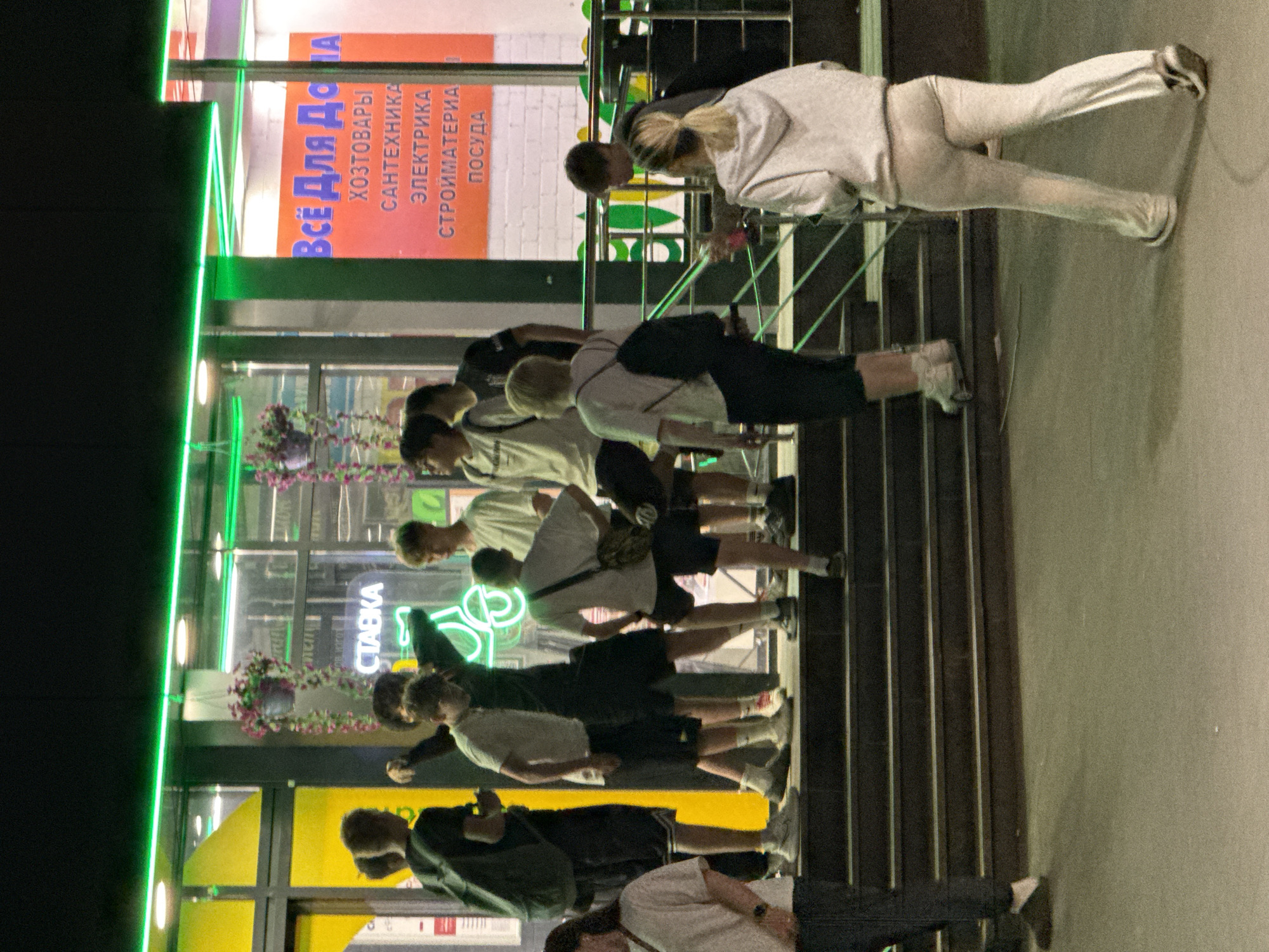The image depicts a nighttime scene outside a commercial building in what appears to be Moscow, Russia, based on the Cyrillic signage. A group of approximately 12 to 15 young individuals, predominantly male, are gathered on and around a set of steps leading up to a brightly lit storefront.
The building facade features large glass windows illuminated by green neon strip lighting above and around the displays. Hanging baskets with purple and pink flowers are suspended near the windows. Visible signs include "Всё Для Дома" (All for Home) on an orange background, listing categories such as "Хозтовары" (Household goods), "Сантехника" (Plumbing), "Электрика" (Electrical), "Стройматериалы" (Building materials), and "Посуда" (Dishware). Another sign displays "СТАВКА" (BET/RATE) in prominent green lettering, accompanied by a green neon outline of a bicycle or scooter, suggesting a betting establishment or similar venue.
The people are dressed in casual attire, including t-shirts, shorts, hoodies, and sneakers. A central group of young men stands on the dark-colored steps; one individual in a white t-shirt and dark shorts gestures with his left arm while looking towards another. Another individual on the steps wears a white t-shirt and carries a camouflage-patterned shoulder bag, while a person further left wears a dark backpack. On the ground level to the right, a young woman in a light-colored tracksuit walks away from the building. To the left, a woman in a white t-shirt and dark pants is partially visible, also in motion. The individuals appear to be socializing or waiting, forming a loose gathering around the entrance. The ground level is a dark, flat surface.