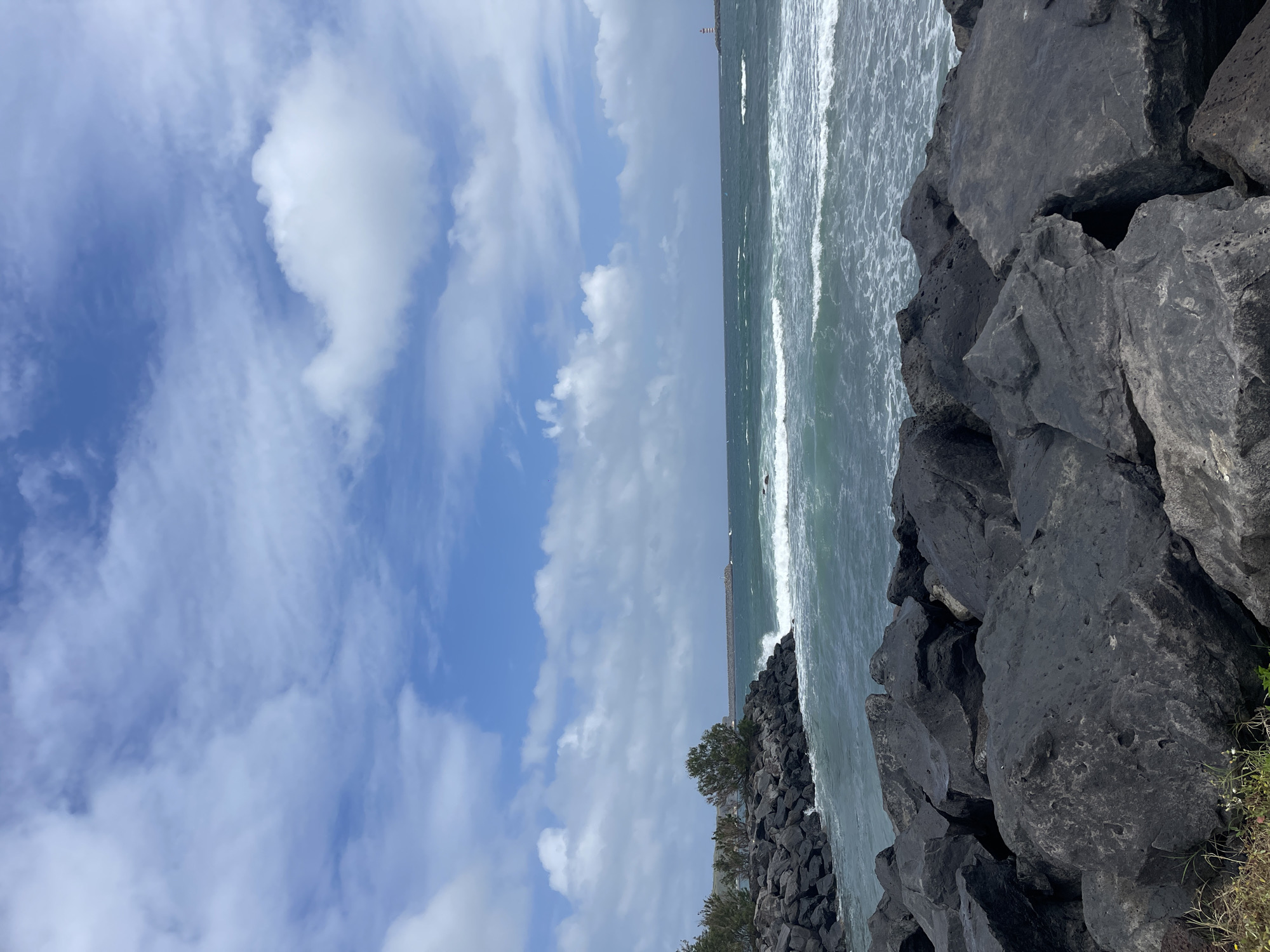 The image captures a coastal scene in Praia da Vitória, Portugal, on a partly cloudy day. The composition is dominated by the view of the Atlantic Ocean meeting a rocky shoreline. Large, dark volcanic rocks form a barrier along the coast, with small green shrubs interspersed among them. The ocean displays moderate waves crashing against the rocks. A breakwater structure extends into the sea, noticeable in the distance. The sky above is a mix of blue and white clouds, suggesting variable weather conditions.