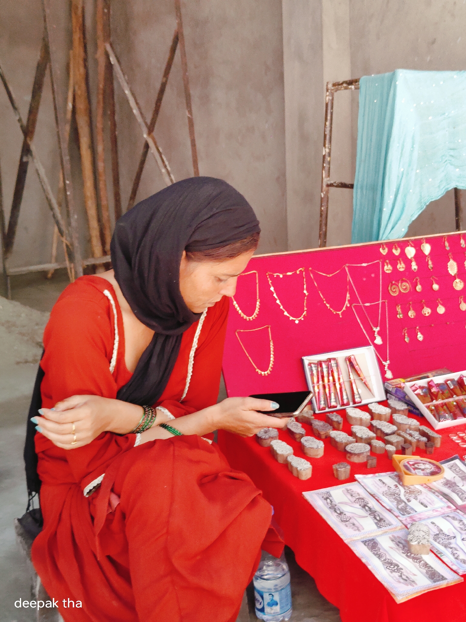 An Indian woman wearing a black hijab and a red outfit sits behind a table displaying various items. She is looking down at a smartphone in her hand. The table is covered with a red cloth and features an assortment of jewelry, including necklaces and earrings, displayed on a pink backing. Below the jewelry, there are several small wooden stamps, possibly for applying temporary tattoos or for printing designs. Boxes containing what appear to be small tools or inks are also visible. In the background, there are metal poles and a draped piece of light blue fabric. The scene appears to be an outdoor market stall, potentially in Leh, India, given the woman's attire and the style of merchandise. A bottle of water is visible on the floor near the table.