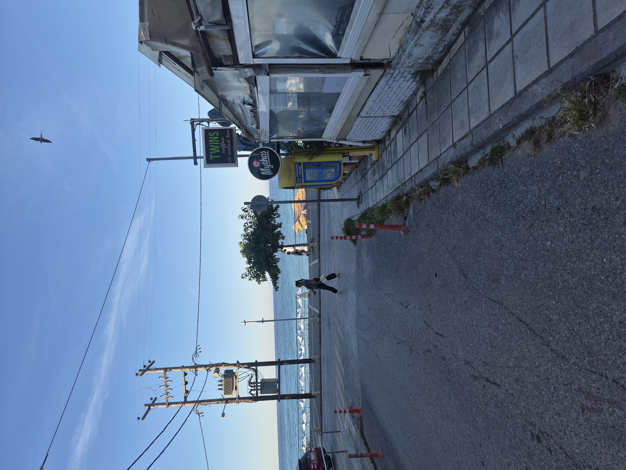 A medium shot captures a street scene in Néa Kallikráteia, Greece, under a clear blue sky. The foreground is dominated by an asphalt road, with tire tracks visible. To the right, a paved sidewalk runs alongside a building with a partially covered exterior, appearing to be a cafe or restaurant. Orange traffic cones are placed along the road and sidewalk. A yellow public telephone booth stands on the sidewalk. Further down the street, a woman is crossing the road. In the background, the blue sea is visible, with rows of white beach umbrellas set up on a beach. A power pole with transformers and electrical wires stands to the left of the road. A sign with "TWINS coffee & salad bar" is mounted on a pole. A bird is seen flying in the sky.