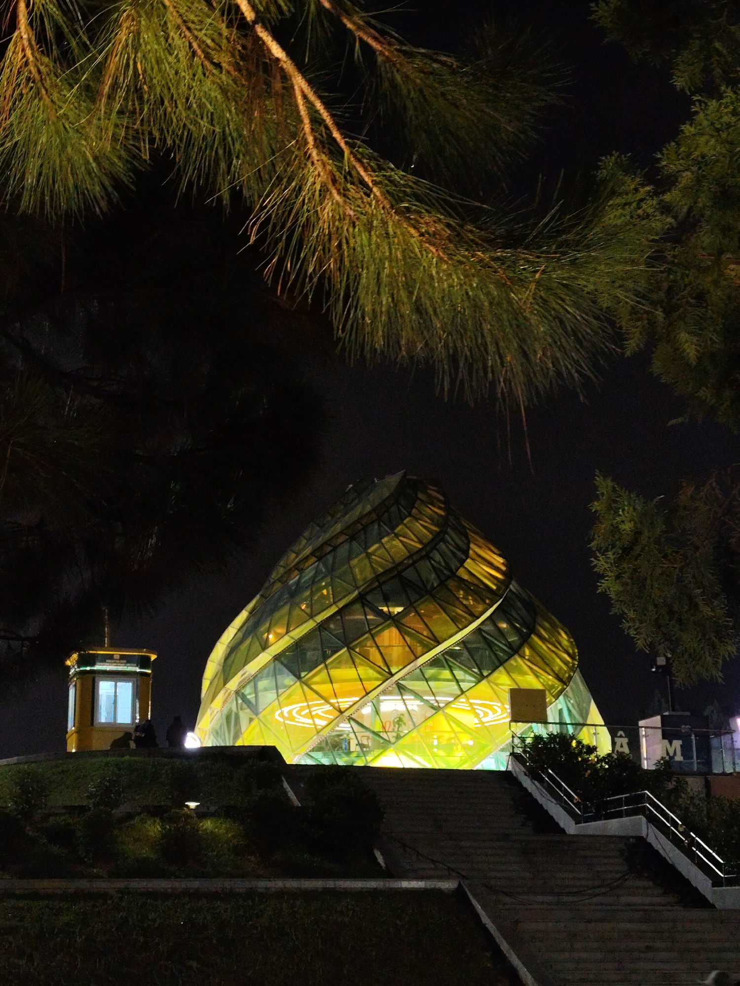 The image is a night shot of a unique building in Da Lat, Vietnam. The building is a spiraling structure made of glass panels, illuminated with a warm, yellow light. A small, yellow booth-like structure sits on the left on a grassy hill, with two silhouetted figures visible on the hill, appearing to be seated. The foreground features a set of concrete stairs leading up to the building. Pine tree branches frame the top and sides of the image. A sign partially visible to the right of the building displays letters "AM" on it. The scene is dark overall, with the building being the main source of light.