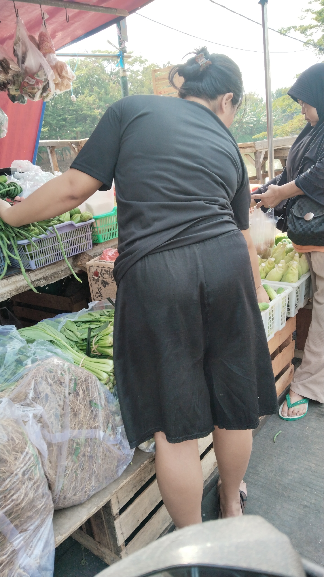 In West Jakarta, Indonesia, an outdoor market scene unfolds. A woman, viewed from the back, is dressed in a black t-shirt and black skirt. She is reaching into a purple plastic basket filled with long green beans. Beside her, another woman wearing a black hijab and a black jacket with a tan shoulder bag is handling produce. Crates and baskets overflow with various vegetables, including what appear to be long beans, leafy greens, and corn. A tarp, possibly serving as a makeshift roof, is visible overhead, and beyond the market stalls, trees and foliage suggest a natural backdrop. The ground appears to be paved.