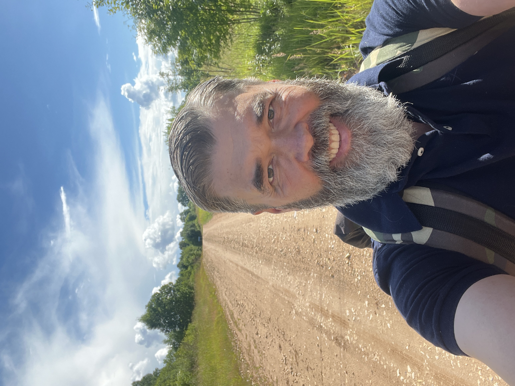 A man with a gray beard smiles at the camera as he stands on a dirt road. He is wearing a dark blue polo shirt and a camouflage backpack. The road stretches into the distance, bordered by grassy fields and trees. The sky is blue with white clouds. The image is taken outdoors in a rural location.