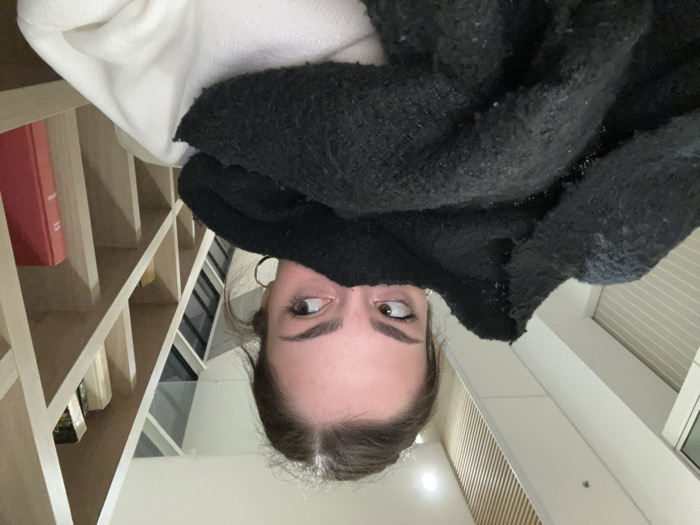 This image, taken in Mundolsheim, France, captures a close-up, low-angle perspective of a young person peeking out from under a dark, textured garment, possibly a sweater or blanket. Only her forehead, eyes, and parts of her cheeks and chin are visible. She has wide, brown eyes and is looking upwards and to her left (the viewer's right if the image were correctly oriented), conveying a curious, playful, or surprised expression. She wears gold hoop earrings.
The background reveals an indoor setting. To the left of the subject, a light-colored wooden bookshelf or shelving unit is visible, partially filled with books, one of which is a prominent red-orange volume. Above the subject's head, a white, possibly fuzzy, garment or blanket is draped. To the right, a window or glass panel with horizontal blinds is visible, set within clean white walls, suggesting a modern interior. A subtle light fixture can be seen in the lower left corner. The lighting indicates it could be daytime or early evening. No legible text is visible within the frame.