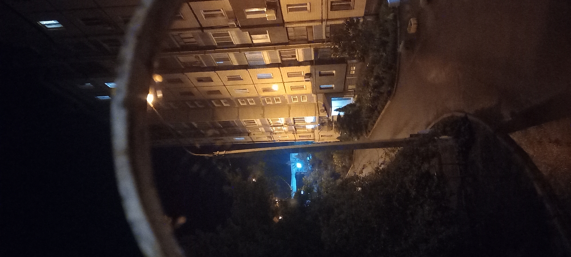 The image captures a nighttime scene in the city of Kryvyi Rih, Ukraine. A multi-story apartment building, likely a Soviet-era design with a brick facade and numerous windows, dominates the right side of the frame. Some windows are illuminated, suggesting occupancy. A street lamp casts a warm yellow glow on the building and the wet asphalt road in front of it, indicating recent rain or dampness. To the left of the building, a dense cluster of dark foliage and trees obscures the ground and part of the building's lower levels. A street runs diagonally across the scene, curving away from the building and disappearing into the darkness. In the background, beyond the trees and across the road, a bright blue light emanates from what appears to be another building or structure, adding a contrasting cool tone to the otherwise warm lighting. The foreground is partially obscured by a blurry, curved, dark object, possibly a railing or a window frame, which creates a frame-within-a-frame effect. The overall atmosphere is subdued and nocturnal, with limited visibility due to the darkness and the focus on artificial light sources.