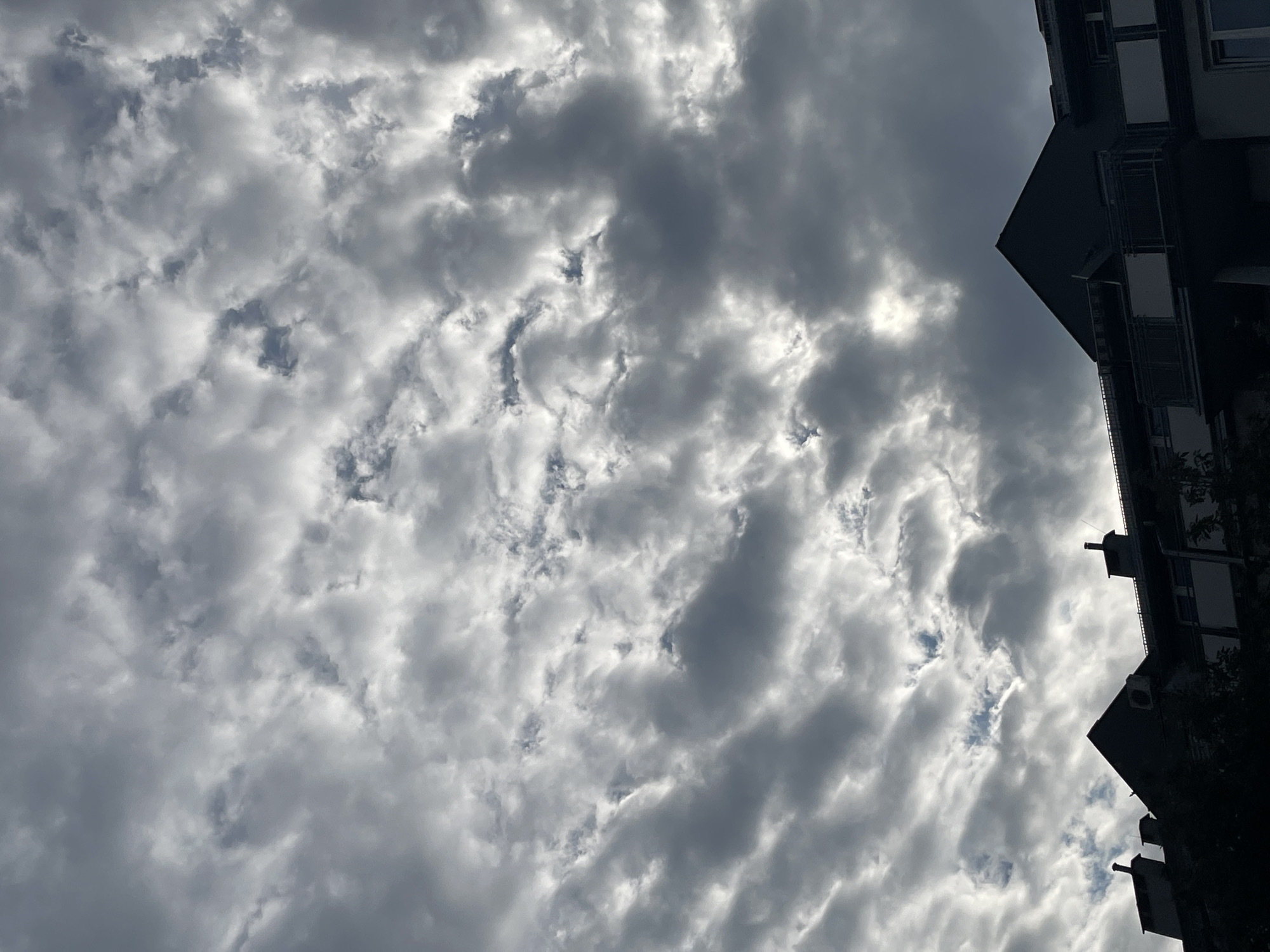 The image depicts a sky filled with altocumulus clouds, partially obscured by a building on the right side. The clouds are mostly light gray with some darker patches, suggesting varying thicknesses. The building is a multi-story structure with visible windows and balconies. The overall scene conveys an overcast day in an urban environment.