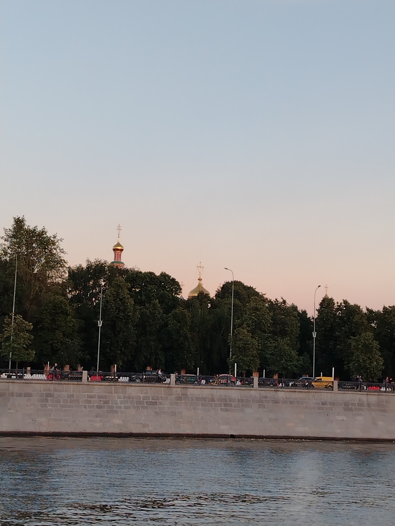 The image depicts a serene riverfront scene in Moscow, Russia, likely during the late afternoon or early evening. The sky is a soft gradient of pale blue and warm pink, suggesting the setting sun. Dominating the mid-ground is a lush line of trees that partially obscures a row of buildings topped with golden domes, characteristic of Russian Orthodox churches. Two distinct church domes with crosses are visible, one more prominent than the other.
In the foreground, a wide, calm river reflects the muted colors of the sky. A stone embankment runs along the river's edge, constructed from large, light-colored rectangular blocks. Along the top of the embankment, a decorative black metal fence encloses a walkway. Beyond the fence, a line of vehicles is visible, including a distinctive yellow taxi. A few scattered figures can be seen walking or standing near the fence, but their details are indistinct. Several streetlights stand tall along the embankment. The overall atmosphere is peaceful and tranquil, capturing a quiet moment by the Moscow River.