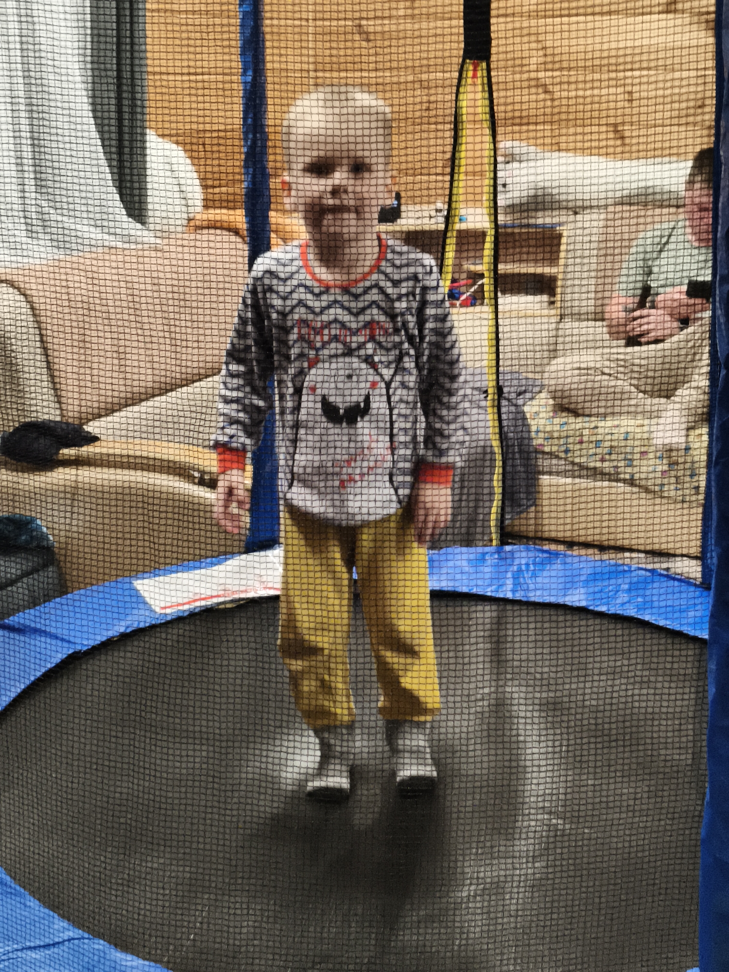 A young child stands on a trampoline inside a room in Русский Вожой, Russia. The trampoline has a blue safety pad and a black jumping surface, surrounded by a black netting enclosure. The child wears a gray, white, and red long-sleeved shirt with a cartoon design and yellow pants with gray socks. Behind the netting, a living room area is visible, featuring a tan couch, wooden walls, and another adult sitting on the couch, using a smartphone.