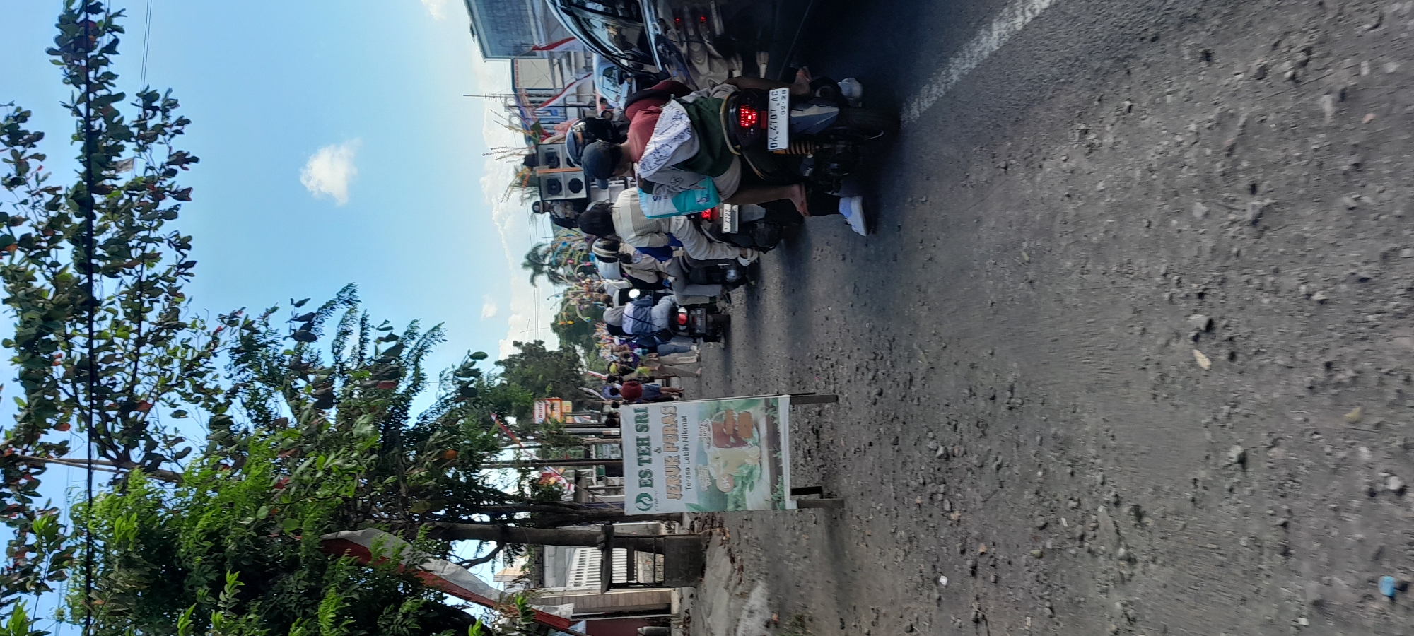 The image depicts a street scene in Rogojampi, Indonesia, characterized by a traffic jam. In the foreground, a white A-frame sign advertises "ES TEH SRI JERUK PERAS" (Sri Iced Tea Squeezed Orange) and states "Terasa Lebih Nikmat" (Tastes Better). The sign features an illustration of an iced tea drink with oranges. A dark-colored car is partially visible on the right side of the image, and numerous motorcycles with riders are stopped in the street, suggesting a traffic standstill. People are seated on the motorcycles, and some are standing on the roadside. Trees with green foliage line the left side of the street, with red and white Indonesian flags or banners draped among the branches. The sky is blue with a few scattered clouds, and the road surface is rough and appears to be asphalt mixed with gravel. The overall impression is a busy, typical day in a bustling Indonesian town.