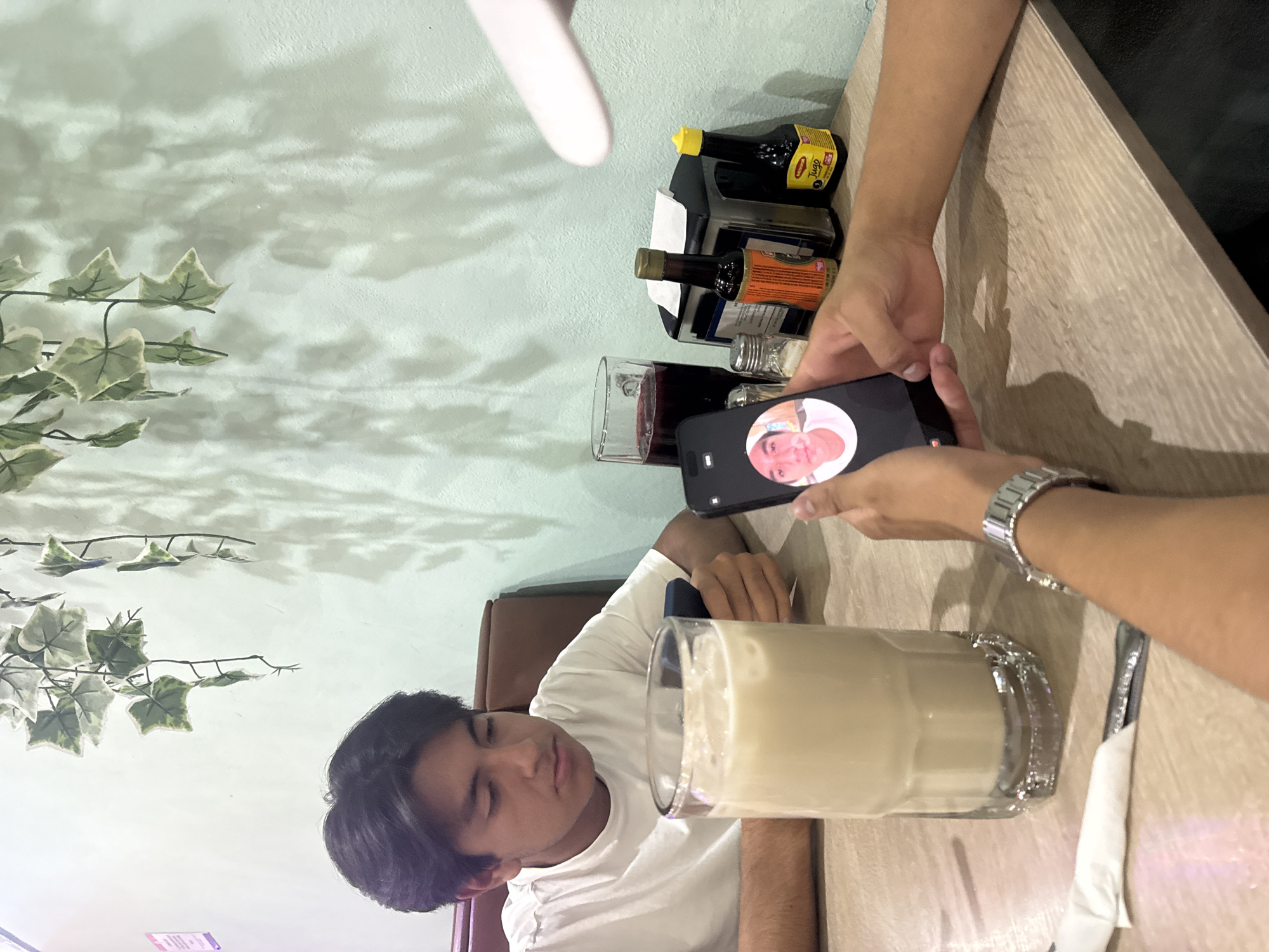 A young man sits at a table in San Bernardino Tlaxcalancingo, Mexico, next to a glass of light brown liquid. Another person's hands hold a cell phone displaying a face. On the table are a glass of red liquid, soy sauce bottles, and silverware. The wall features ivy-like decorations.