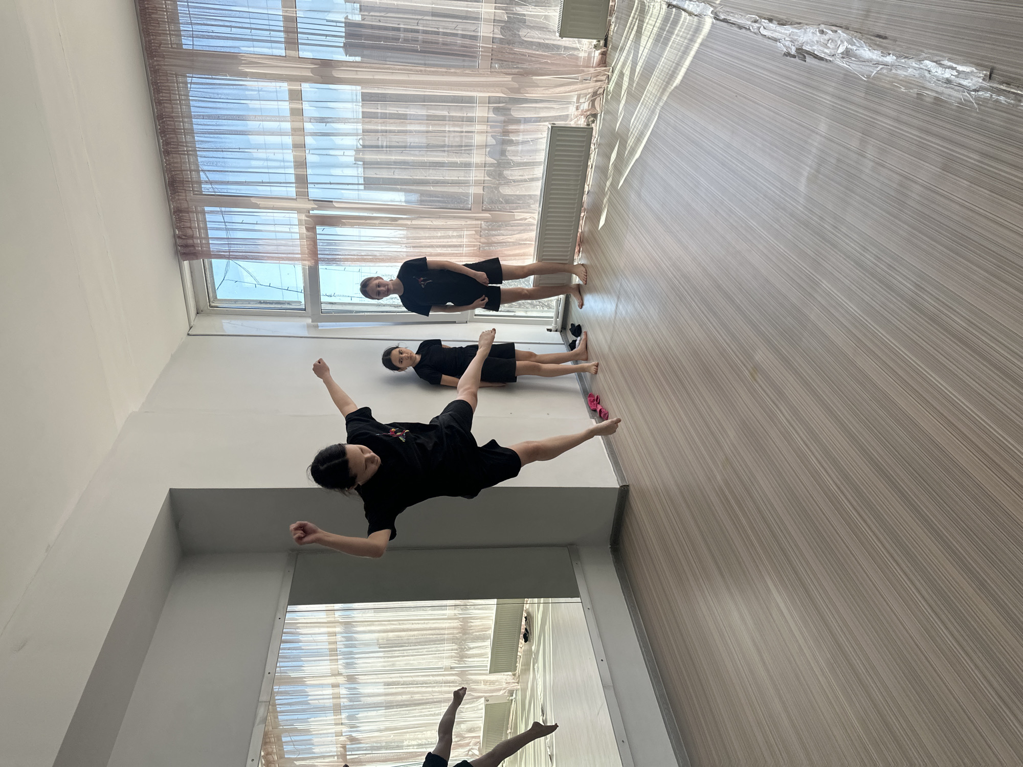 The image shows the interior of what appears to be a dance or exercise studio. There are three individuals dressed in black t-shirts and shorts. One person is in mid-air, seemingly in a dance pose. Another is lying on the floor, and the third is standing. The studio has large windows with sheer curtains, a white wall, a radiator, and a light-colored wooden floor. A mirror is placed along one of the walls, showing a partial reflection of another person. The location is likely in Votkinsk, Russia, as requested.