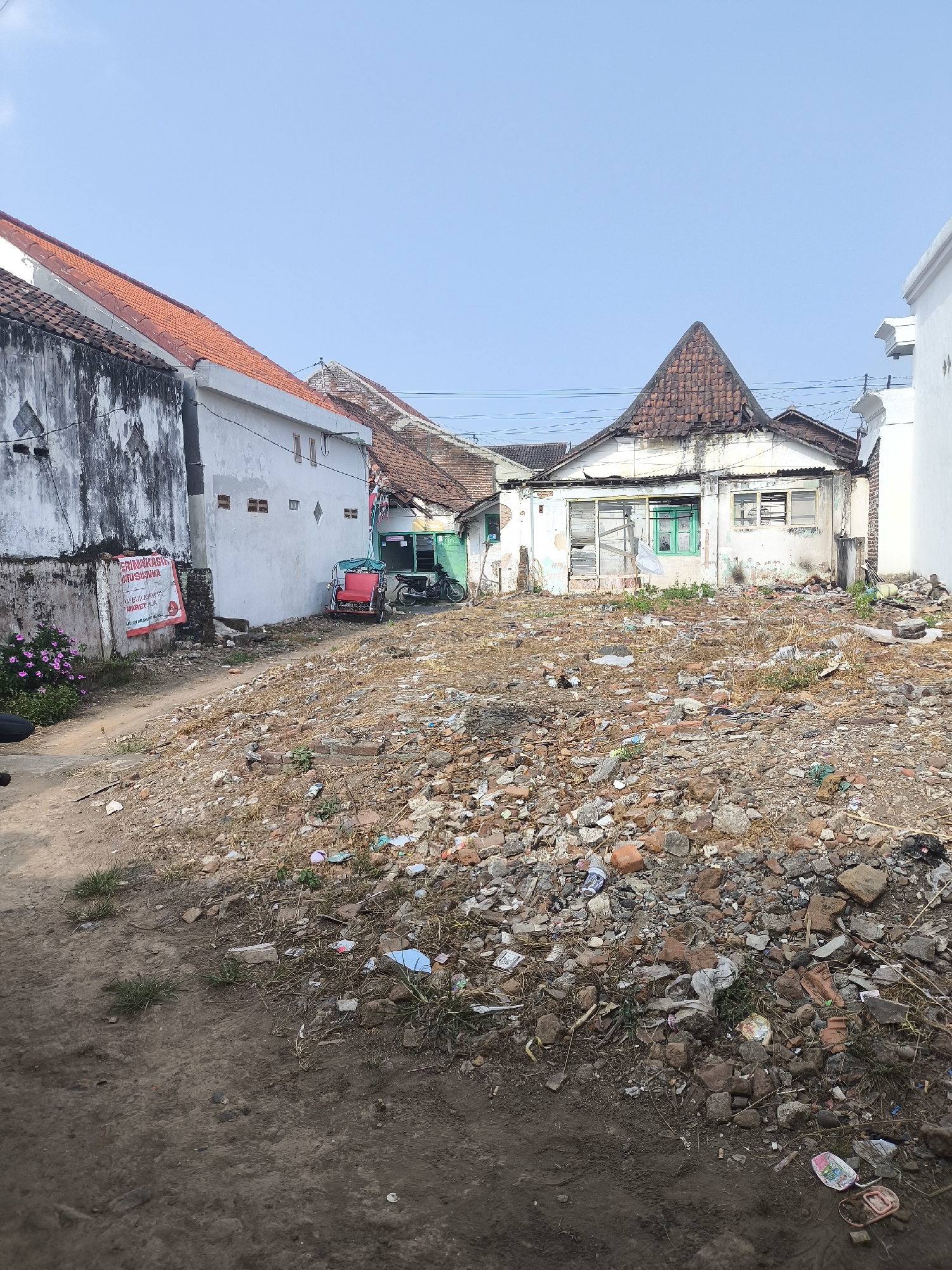 The image depicts a residential area in Lumajang, Indonesia, characterized by a steep, debris-filled slope leading up to several houses. The houses show signs of wear and tear, with some appearing dilapidated. A red and blue rickshaw is parked next to a building with green-framed windows. A motorcycle is parked further back. The slope itself is covered with a mix of soil, rocks, broken bricks, and scattered trash, including plastic bottles and bags. There are some small patches of grass and weeds growing amongst the debris. A poster is attached to a white wall on the left side of the image. The sky is clear and blue.