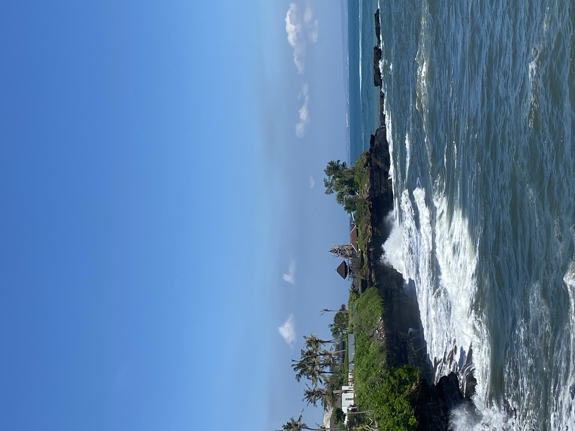 The image depicts a coastal scene in Mengwi, Indonesia. The view features a rocky coastline with waves crashing against the rocks. The vegetation includes greenery and palm trees. The sky is blue with a few scattered clouds. A small structure resembling a temple is visible on the rock formation. The ocean occupies the right side of the image, characterized by its choppy surface and white foam where the waves meet the rocks. The image is oriented vertically.