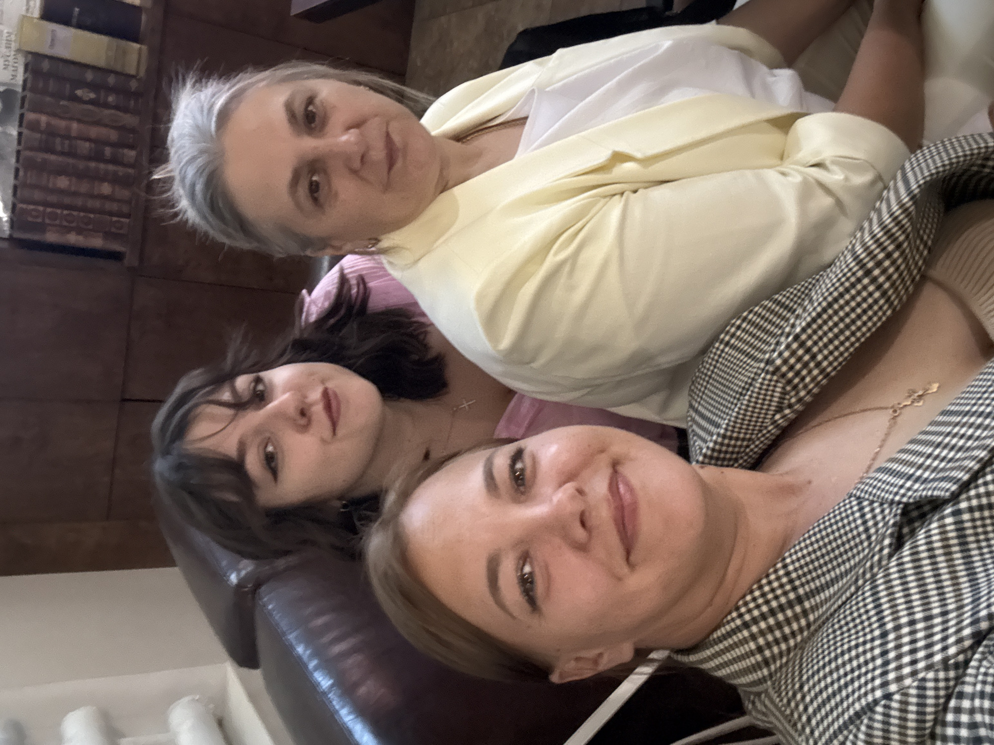 Three individuals, two women and a younger female, are posing for a selfie on a sofa. The woman in the foreground, on the left, wears a black and white checkered blazer and a necklace. To her right, a woman with graying hair wears a light yellow blazer over a white top. Behind them, a younger woman with dark hair and bangs wears a pink top and a cross necklace. The background shows a wooden shelf filled with books and a portion of a white wall. The scene appears to be an indoor setting, possibly a living room or a lounge area in Dmitrov, Russia.