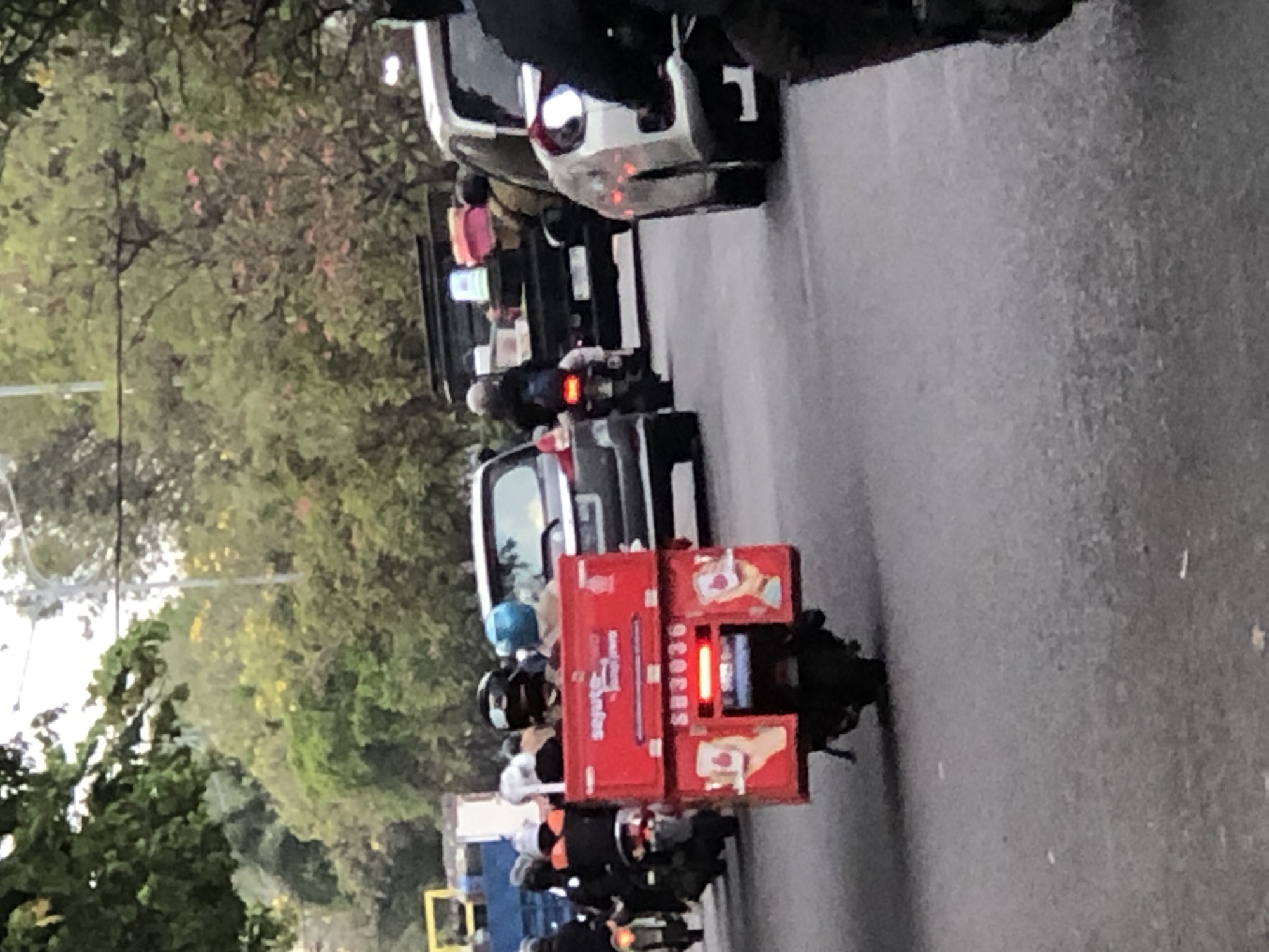The image shows a street scene in Sumorame, Indonesia, with several motorcycles and cars on a paved road. In the foreground, a red delivery box is mounted on the back of a motorcycle. The box has a logo that appears to say "Sajuto" and a license plate that reads "SB3036". Two people are visible riding on the motorcycle, both wearing helmets. Behind this motorcycle, other motorcycles and vehicles are present, suggesting a busy street. Trees and foliage line the sides of the road, with some buildings visible in the background. The sky is overcast, indicating a cloudy day.