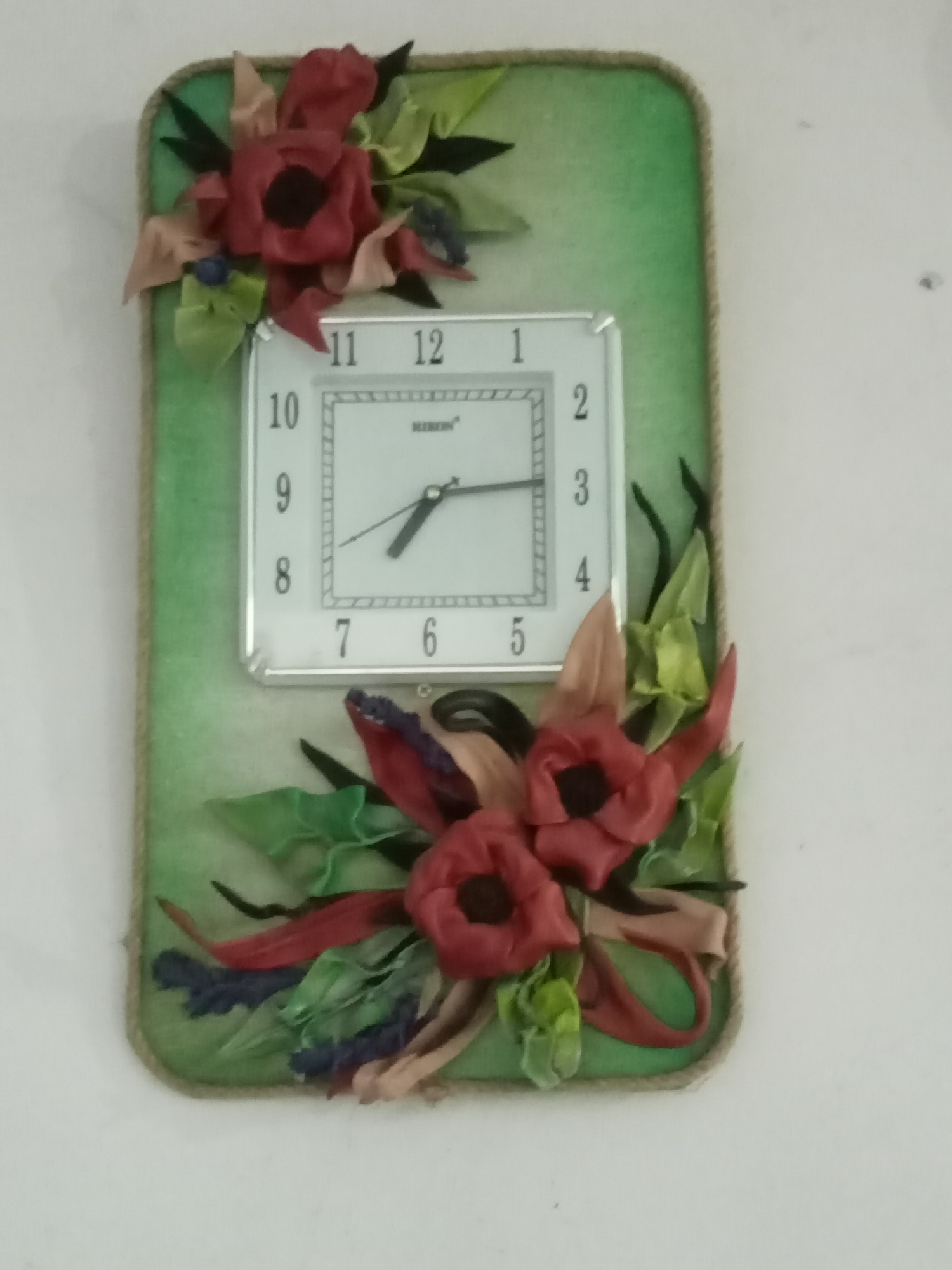 A decorative wall clock with a white square face and black numerals is mounted on a rectangular plaque with a green background. The plaque is edged with rough twine. Two floral arrangements, made of red and pink ribbon flowers with green leaves, are attached to the plaque, one at the top left and one at the bottom center. The clock face has a brand name "REMSO" printed on it. The time displayed on the clock is approximately 10:40. The clock is hanging on a plain white wall.
