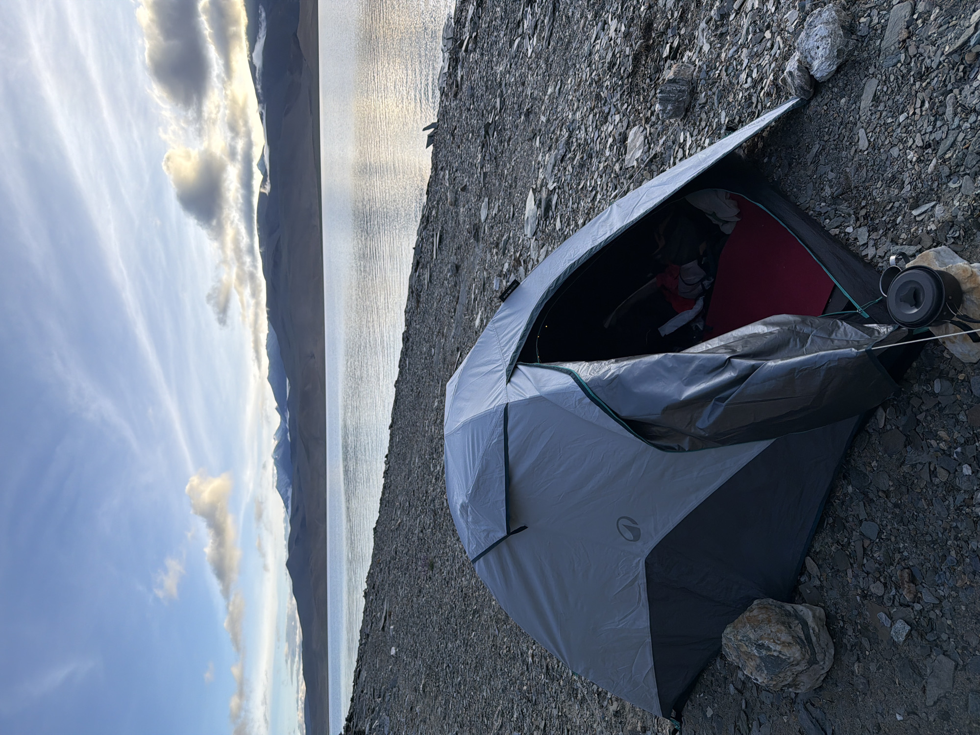 A gray and black tent is pitched on a rocky shore next to a vast, calm body of water. The tent is partially open, revealing a red sleeping pad inside and some belongings. In the foreground, a small black pot with a lid and a mug sit on a rock. The water stretches to the horizon, where it meets a range of distant, dark mountains with snow-capped peaks under a partly cloudy sky. The overall scene depicts a remote, natural landscape, possibly a campsite at high altitude. The location is identified as Unknown, India.