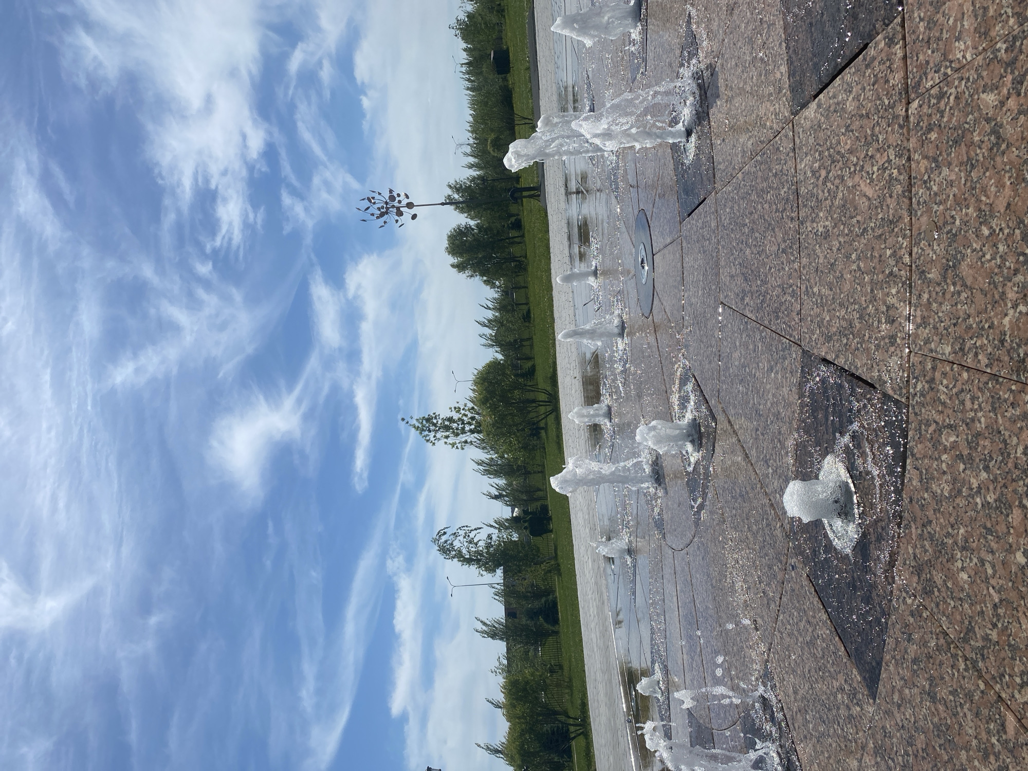 The image captures a vibrant public space in Tyumen, Russia, featuring a ground-level fountain area, lush greenery, and a bright sky. The scene is dominated by a series of pop-up water fountains jetting water upwards from a tiled surface made of mottled brown and black granite-like material. The water creates splashes and wets the surrounding tiles, reflecting the bright sunlight.
In the background, a strip of well-maintained green grass and numerous trees and bushes stretch horizontally, forming a natural barrier. Scattered among the trees are several tall street light poles. To the left of the center, a distinctive decorative sculpture resembling a stylized metal pinwheel or small, sparse tree stands tall against the sky.
The sky is a clear, brilliant blue, adorned with wispy, delicate white clouds that spread across the upper portion of the frame. The overall impression is that of a pleasant, sunny day. The low camera angle emphasizes the upward spray of the fountains and the vastness of the sky. No people are visible in the scene, and there is no discernible text. The setting appears to be a modern park or plaza designed for public enjoyment and recreation.