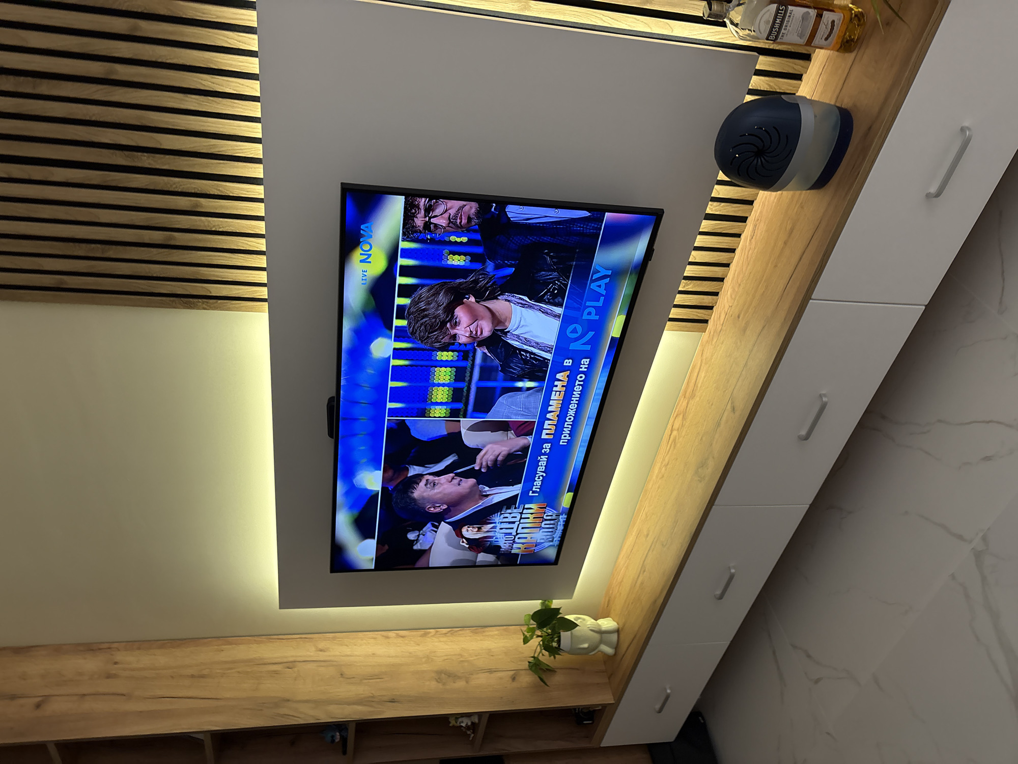The image captures a modern living room interior, likely in Sofia, Bulgaria, featuring a wall-mounted television displaying a live broadcast. The TV is centered on a light grey panel, flanked on the left by a decorative wooden slat wall feature illuminated from behind with warm light.
On the television screen, identified by "LIVE NOVA" in the top left, a show is in progress, possibly a talent competition. Several individuals are visible: a person with brown hair and a dark jacket in the foreground, another person with glasses and a beard behind them, and a man in a white shirt and dark jacket further down, looking upwards. An overlay text on the screen reads "Гласувай за ПЛАМЕНА в приложението на 2 PLAY," which translates to "Vote for PLAMENA in the 2 PLAY application."
Above and to the right of the TV, on a wooden shelf, a bottle of "BUSHmills IRISH WHISKEY ORIGINAL" and a blue and silver air freshener or small humidifier are present. Below the TV, on another wooden shelf, a small green plant in a white, head-shaped decorative pot adds a touch of greenery.
The room's aesthetic is clean and contemporary, combining light wooden tones with white and light grey surfaces. White cabinets with sleek silver handles are visible on the far right, set against a light-colored wall with a subtle marbled pattern. The lighting is warm and ambient, suggesting an indoor setting, likely in the evening.