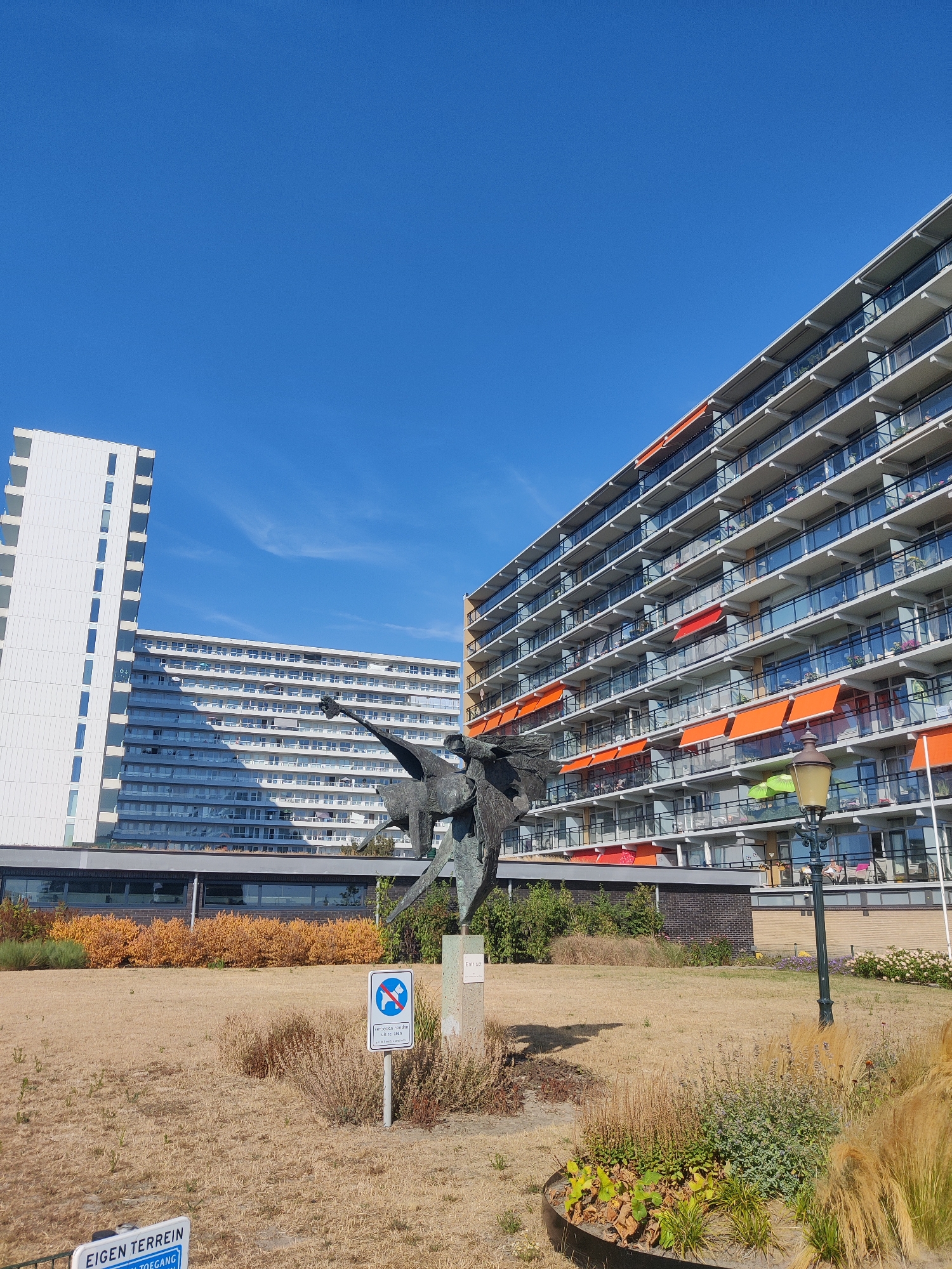This image shows the Sculpture of the Dancing Couple, also known as "Dansend Paar" or "De Dansende Man en Vrouw," located in The Hague, Netherlands. The sculpture is a dark metal abstract piece depicting two figures in a dynamic pose. It is situated in a grassy area with dry, brown grass and some decorative plants.
Behind the sculpture are two large, modern apartment buildings. The building on the right is a multi-story structure with numerous balconies, many of which have orange retractable awnings. The building on the left is a tall, white, rectangular building with a more uniform facade of windows.
In the foreground, there are signs. One sign, partially visible at the bottom left, appears to be a blue and white sign with text that includes "EIGEN TERREIN" and "TOEGANG." To the right of this, a smaller white sign on a pole shows a blue circle with a red diagonal line across a depiction of a dog and a person, indicating a prohibition, likely related to pets.
A lamppost with a dark, ornate design stands to the right of the sculpture and the sign. The sky is a clear, bright blue with no clouds visible. The overall scene suggests a sunny day in an urban or suburban park setting with residential buildings.
