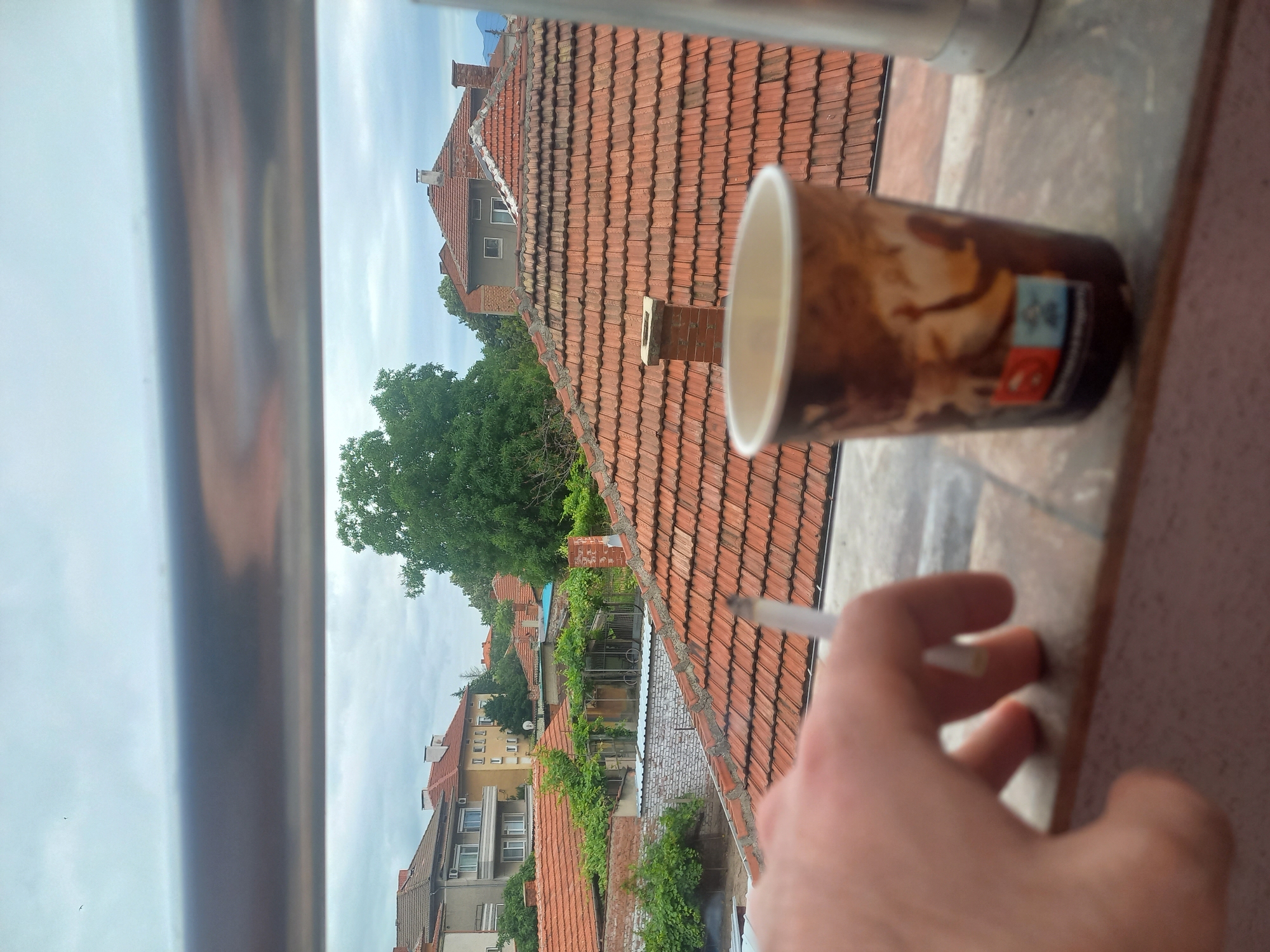 In an outdoor urban setting, a close-up shot frames a hand holding a lit cigarette. To the right, a disposable coffee cup with a busy pattern is positioned on a windowsill. The background reveals a panorama of red-tiled rooftops and buildings under an overcast sky, suggesting a residential area in Parvomay, Bulgaria. The overall atmosphere is casual and contemplative.